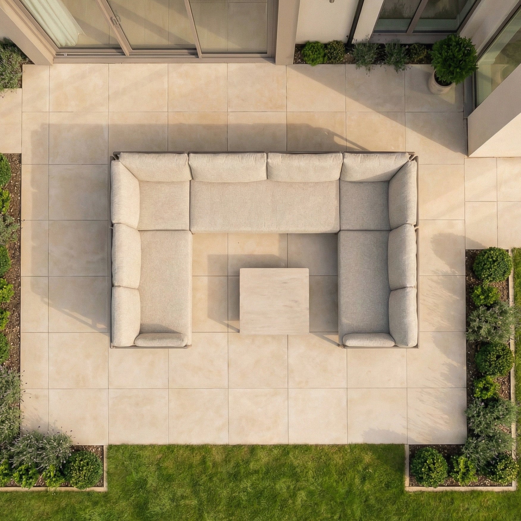 An outdoor patio features the Sorelli U Shape Corner Sofa Set in Oatmeal and a square table, surrounded by greenery and viewed from above.