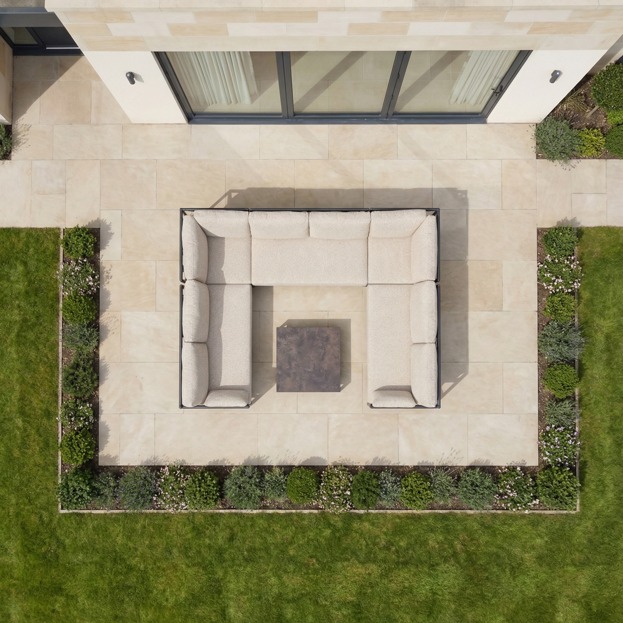 Aerial view of a patio showcasing the Sorelli U Shape Corner Sofa Set in Truffle arranged around a square table, ideal for outdoor entertaining and surrounded by garden furniture, grass, and shrubs.