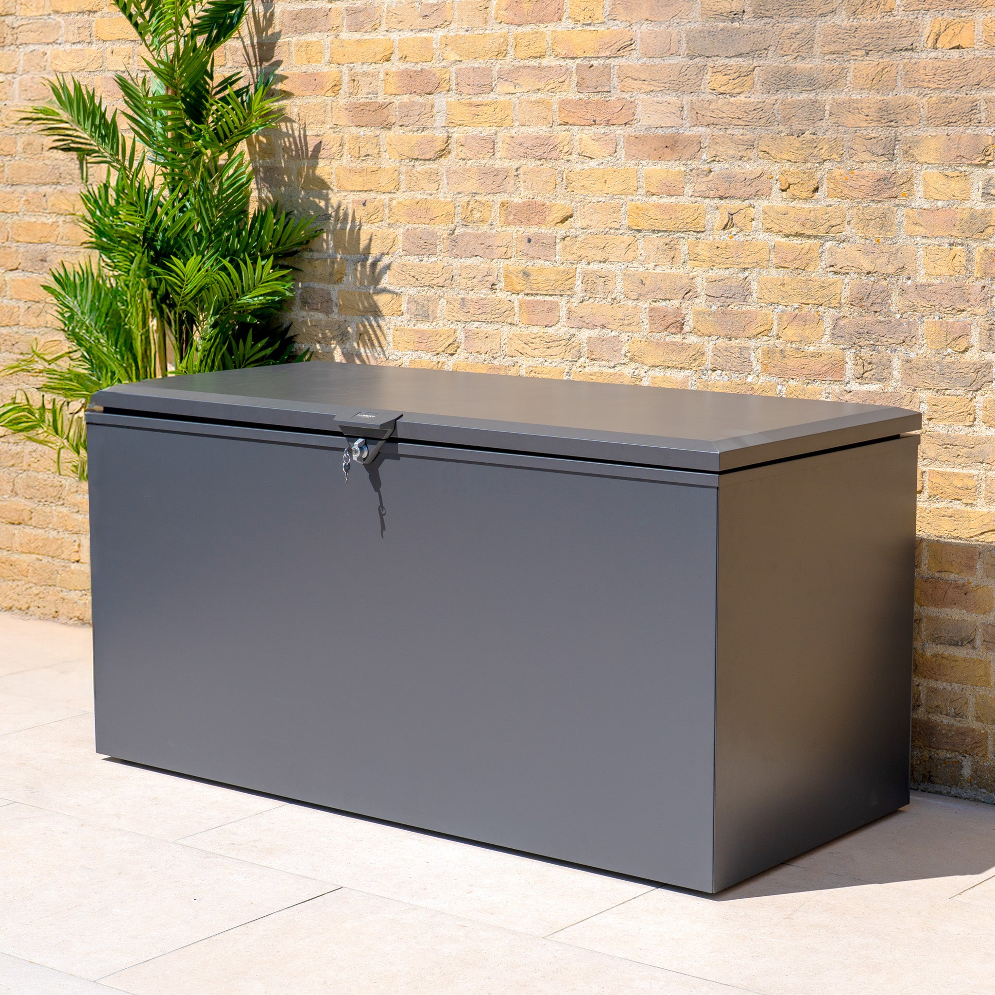 The Large Steel Storage Box in Grey sits on the patio by a brick wall and green plant, providing secure, watertight outdoor storage.