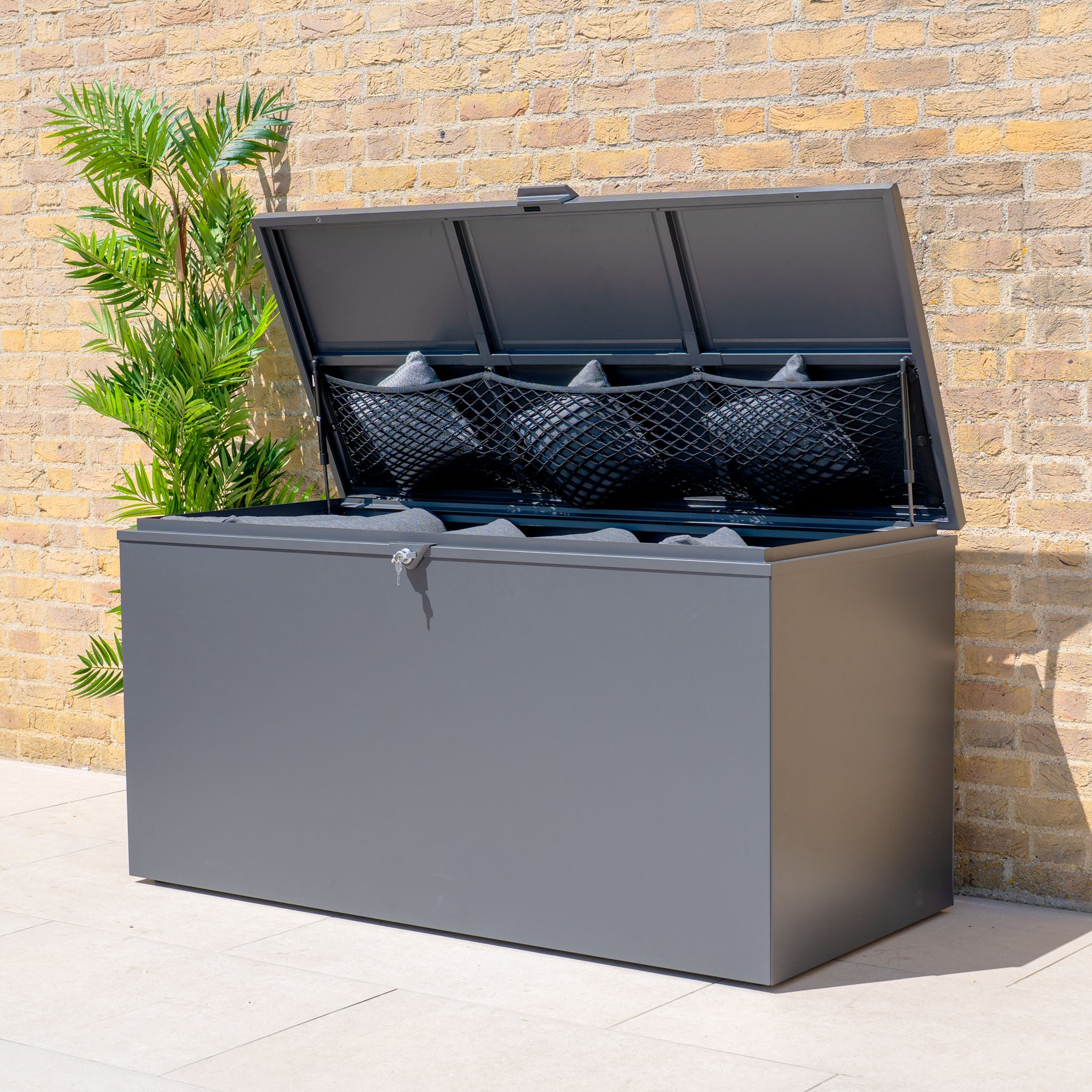 The Large Steel Storage Box in Grey is shown with its lid open, displaying black cushions and a net pocket inside. The box features a watertight seal for outdoor use.
