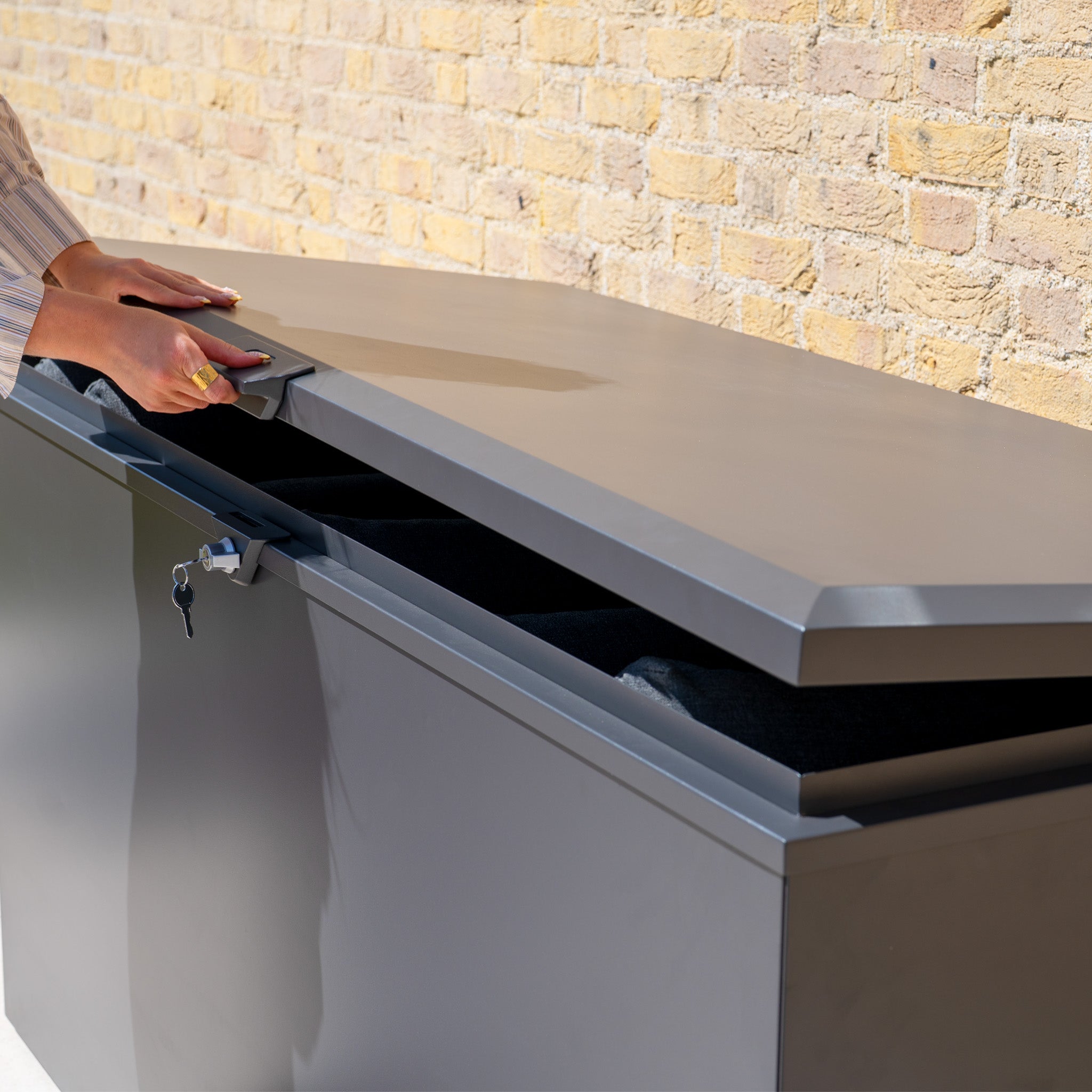A person unlocks a Large Steel Storage Box in Grey with a key; the watertight, outdoor-ready box sits against a brick wall.