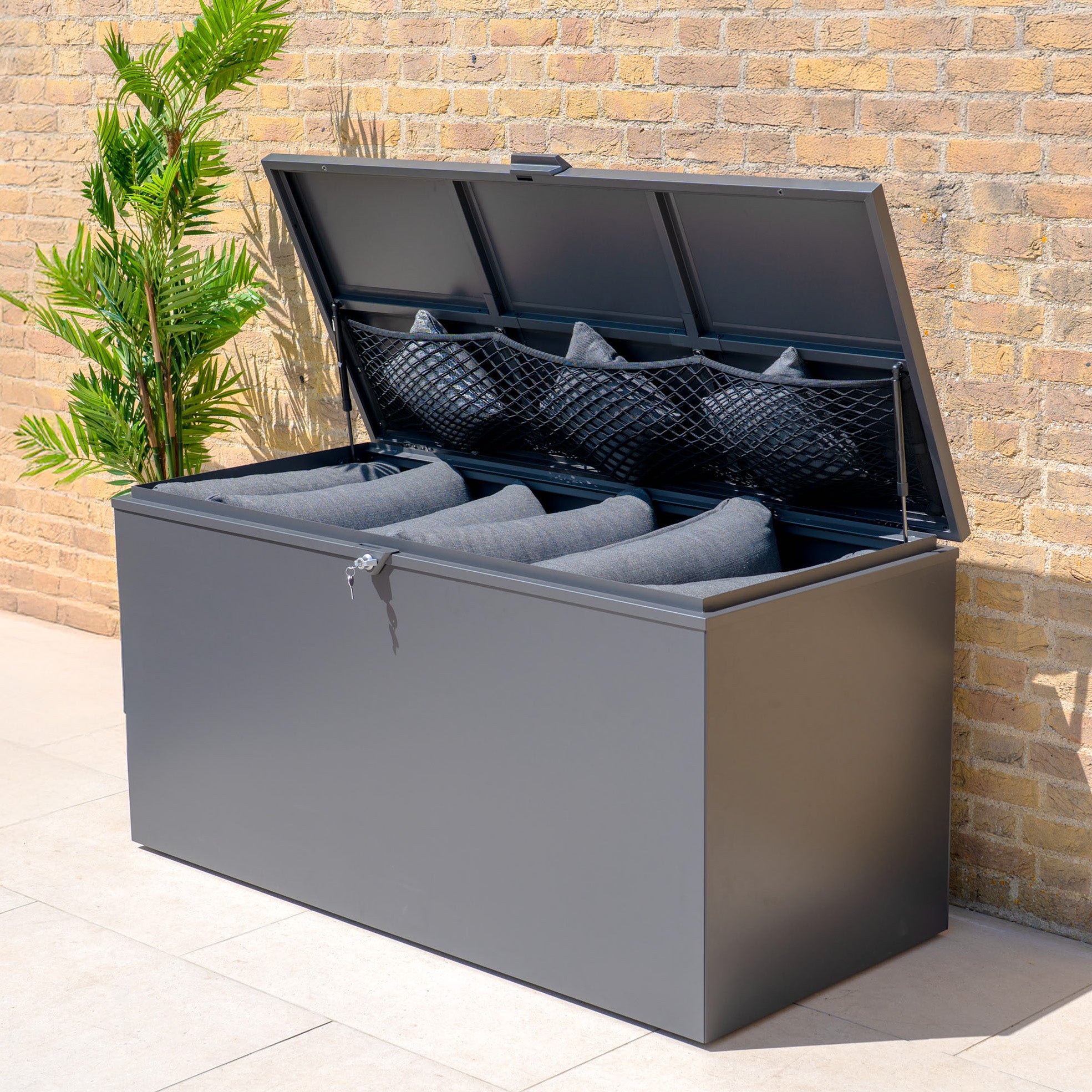 The Large Steel Storage Box in Grey with a watertight seal keeps patio essentials protected and organized. Its lifted lid reveals spacious outdoor storage for gray cushions, blending perfectly with your decor by the brick wall and plant.