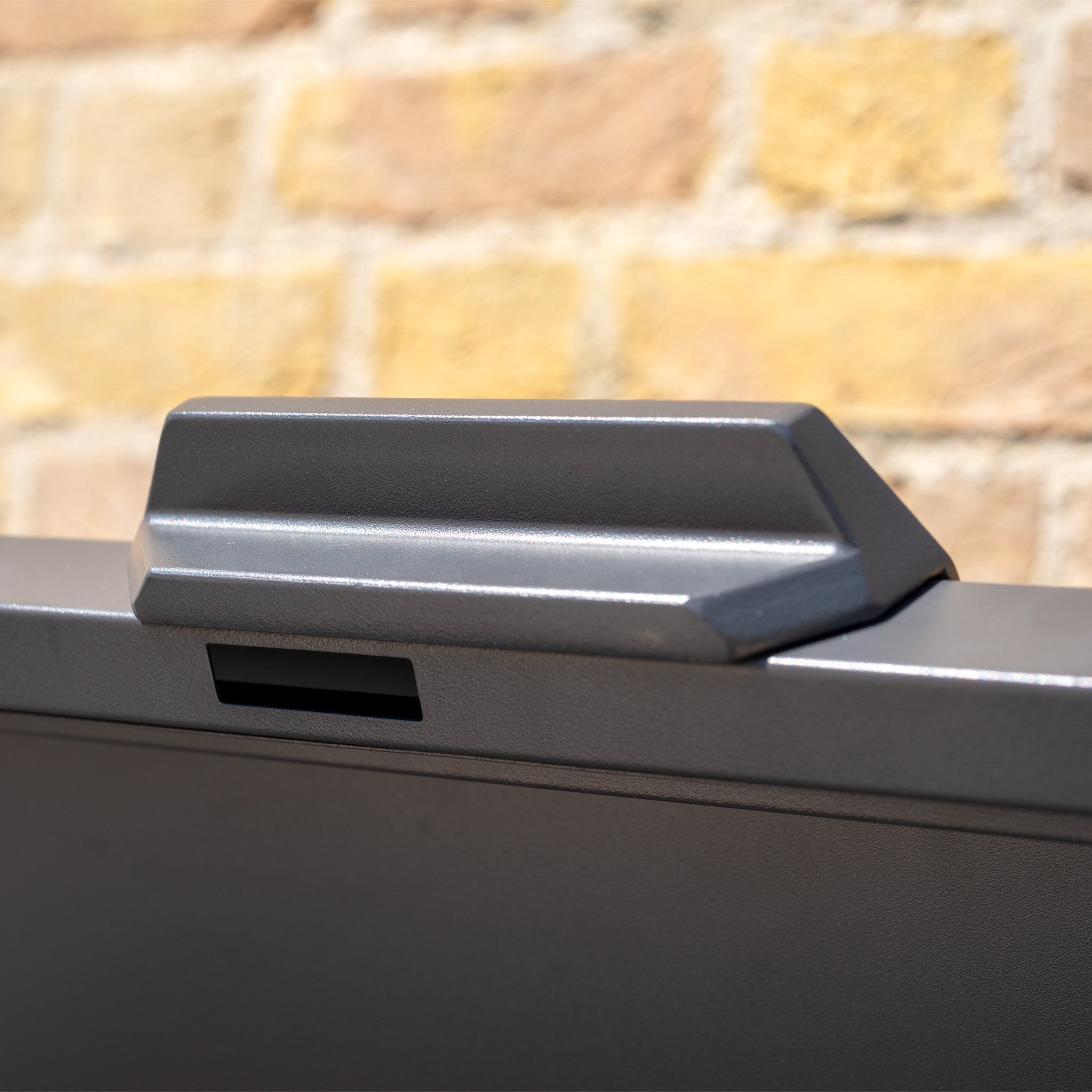 Close-up of the gray handle on a Large Steel Storage Box in Grey, featuring a watertight seal, placed on a dark surface with a blurred brick wall in the background.
