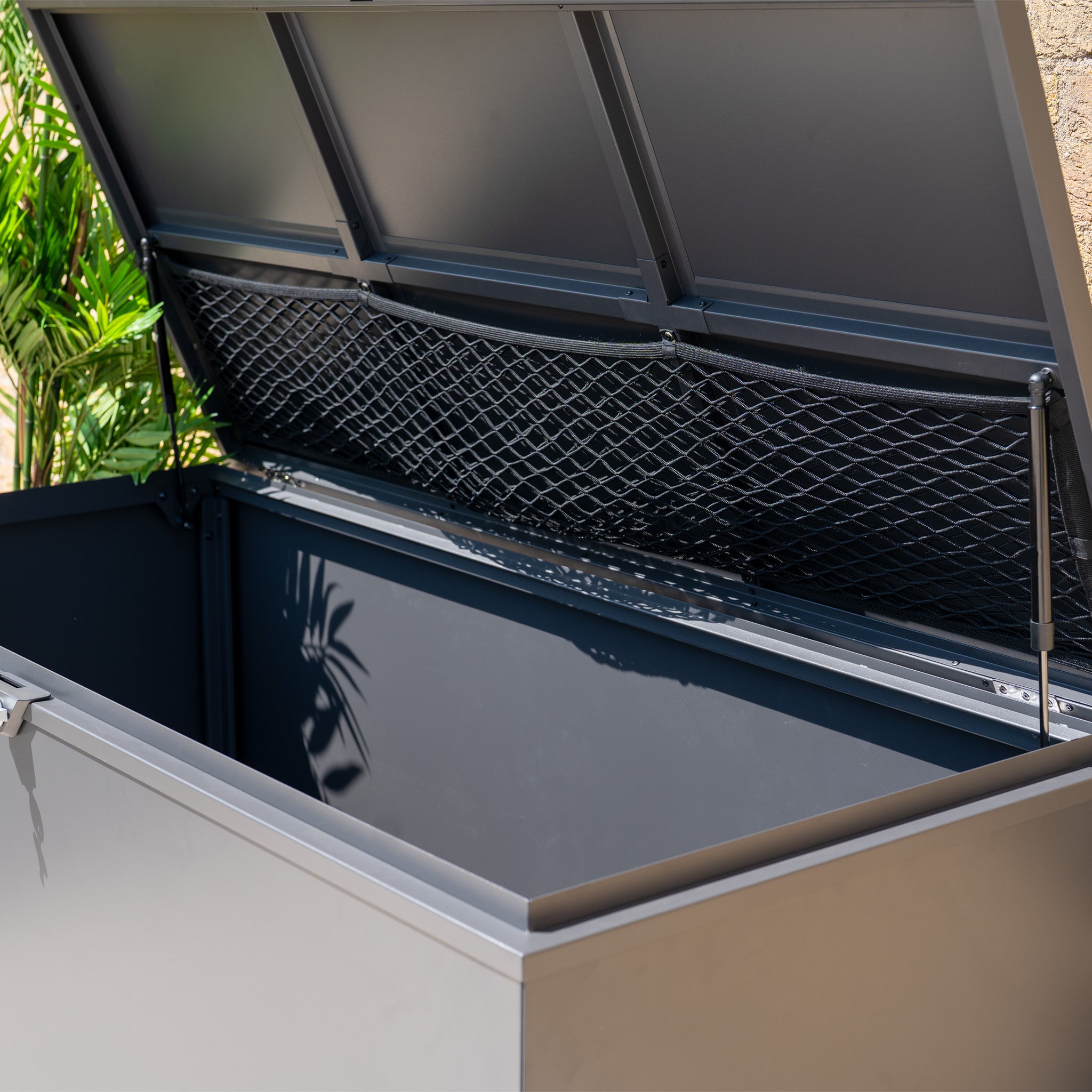 The Large Steel Storage Box in Grey features a watertight seal, spacious open design, and interior net pocket—perfect for outdoor storage. Lush plants can be seen in the background.