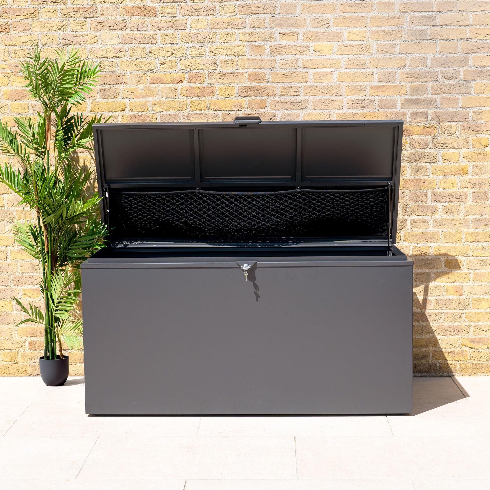 The Large Steel Storage Box in Grey, with a watertight seal and open lid, sits by a brick wall and potted plant—ideal for secure outdoor storage.