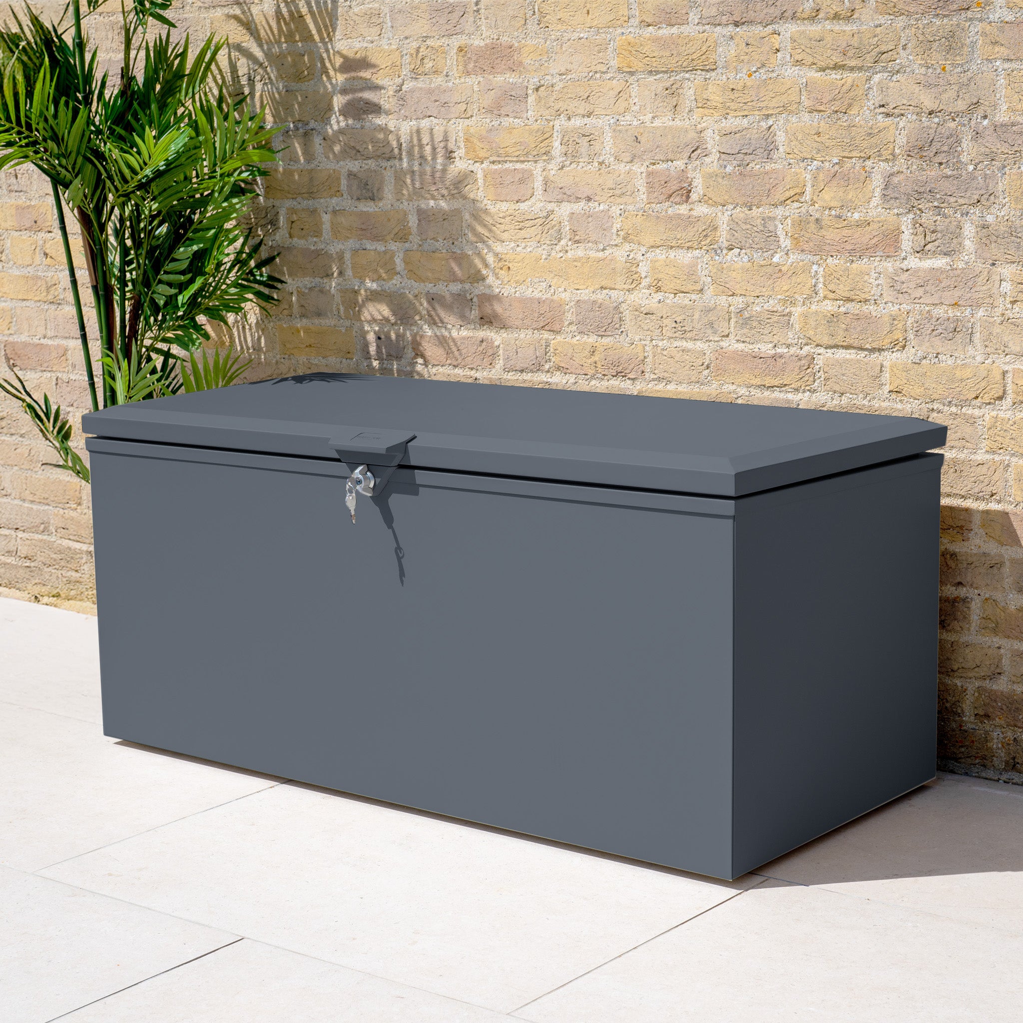 The Small Steel Storage Box in Grey sits on a patio against a brick wall near a green plant—ideal as a waterproof garden storage solution to keep your items safe and dry.