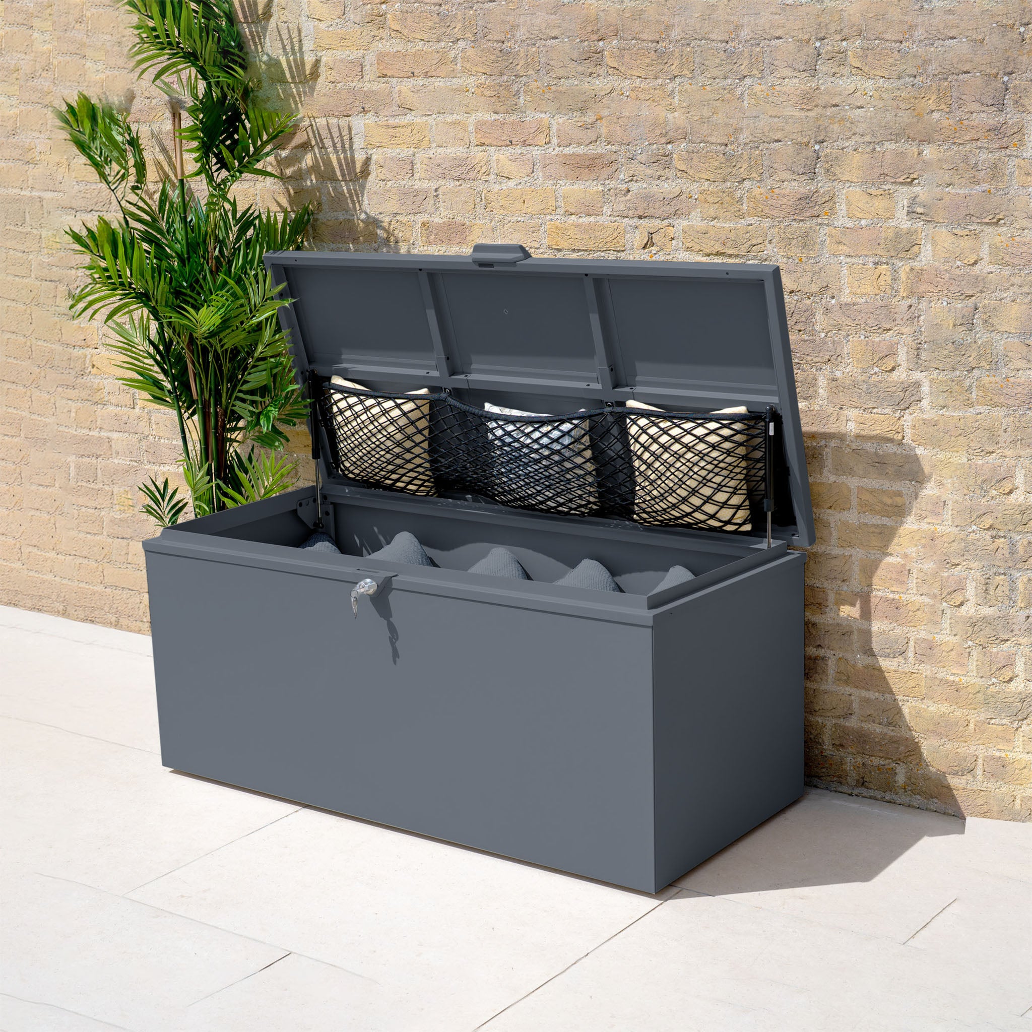 The Small Steel Storage Box in Grey, shown with its open lid holding wicker baskets, is placed beside a brick wall and a potted plant.