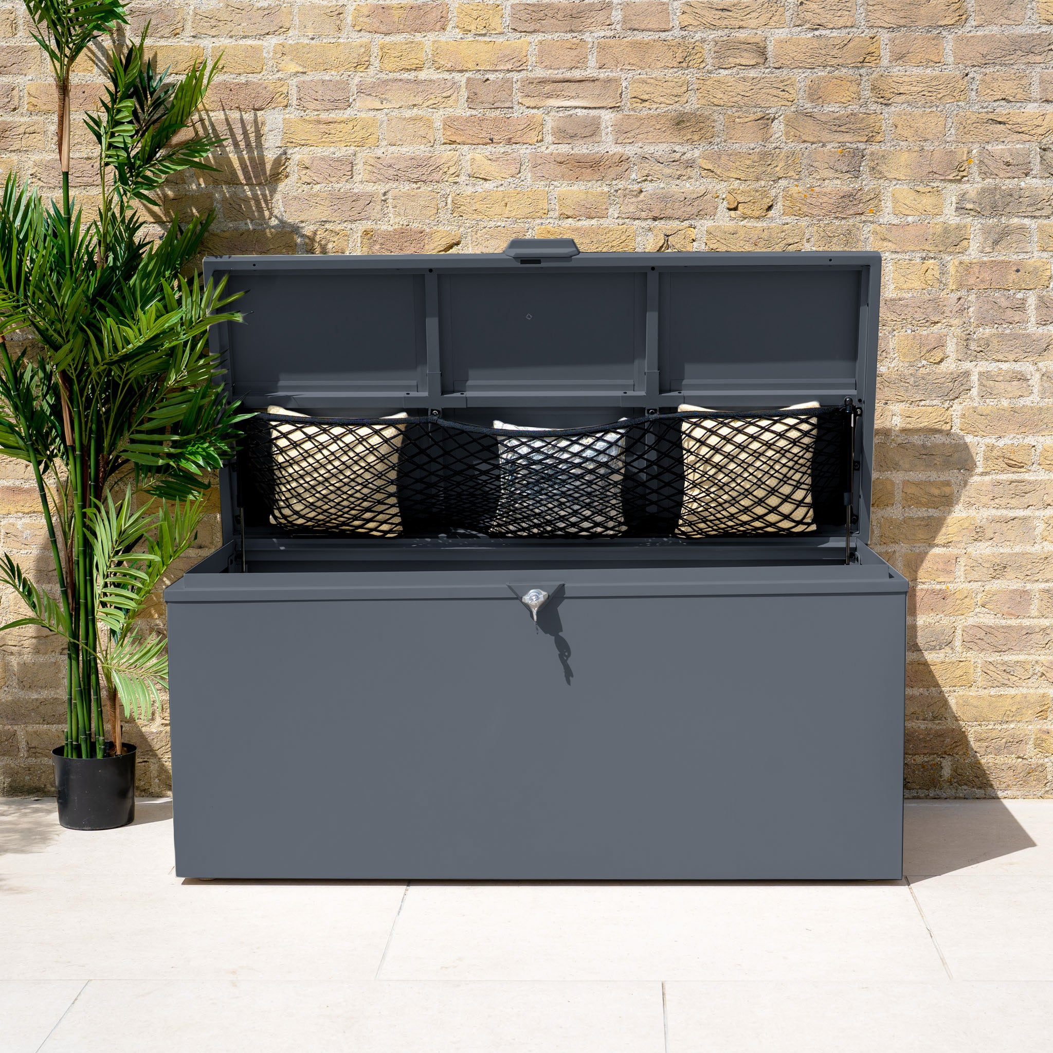 The Small Steel Storage Box in Grey sits against a brick wall, its open lid revealing cushions and a mesh net inside.