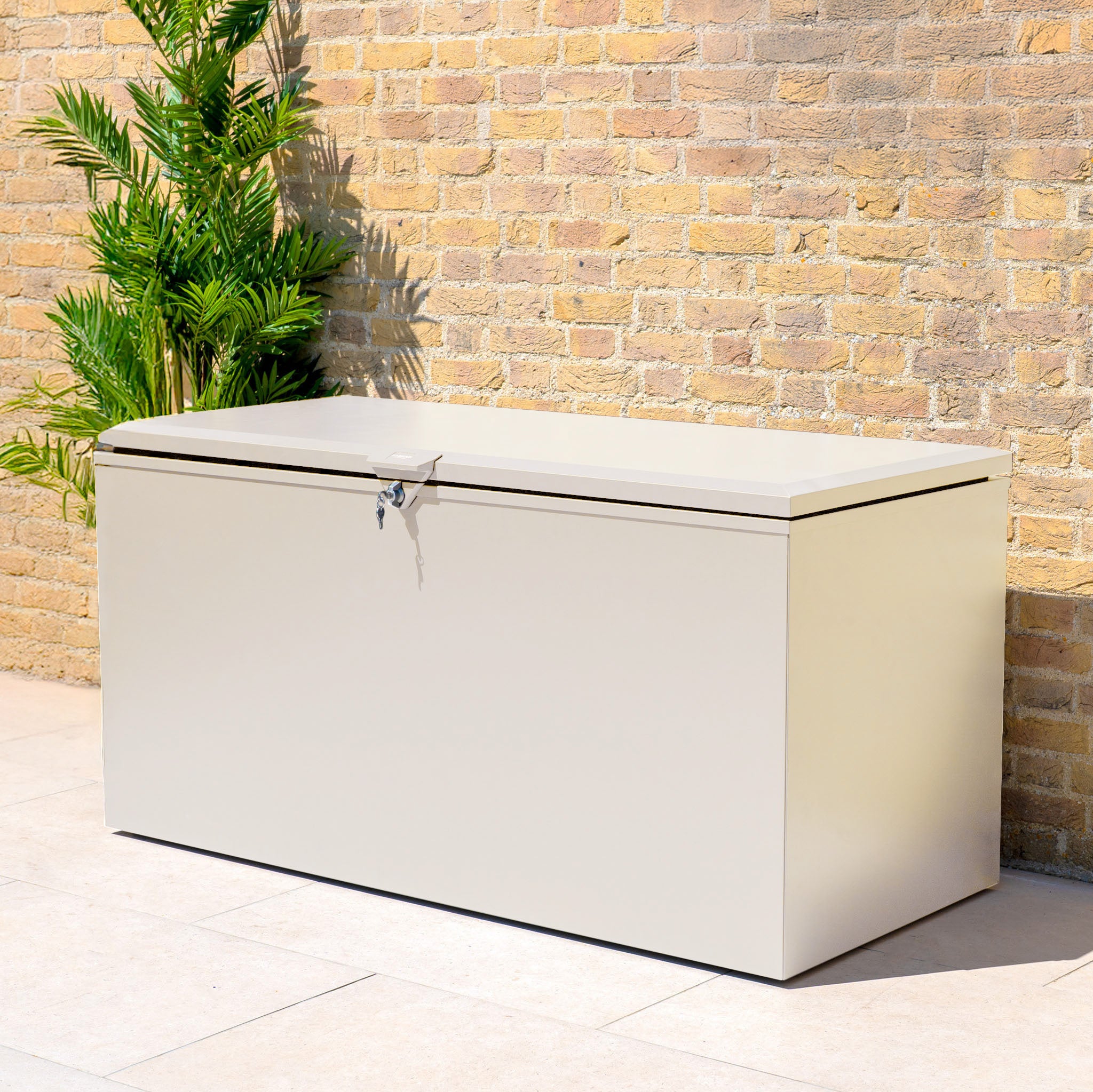 The Large Steel Storage Box in Latte, featuring a watertight seal and lock, sits against a brick wall near a green plant to keep your items safe and dry outdoors.