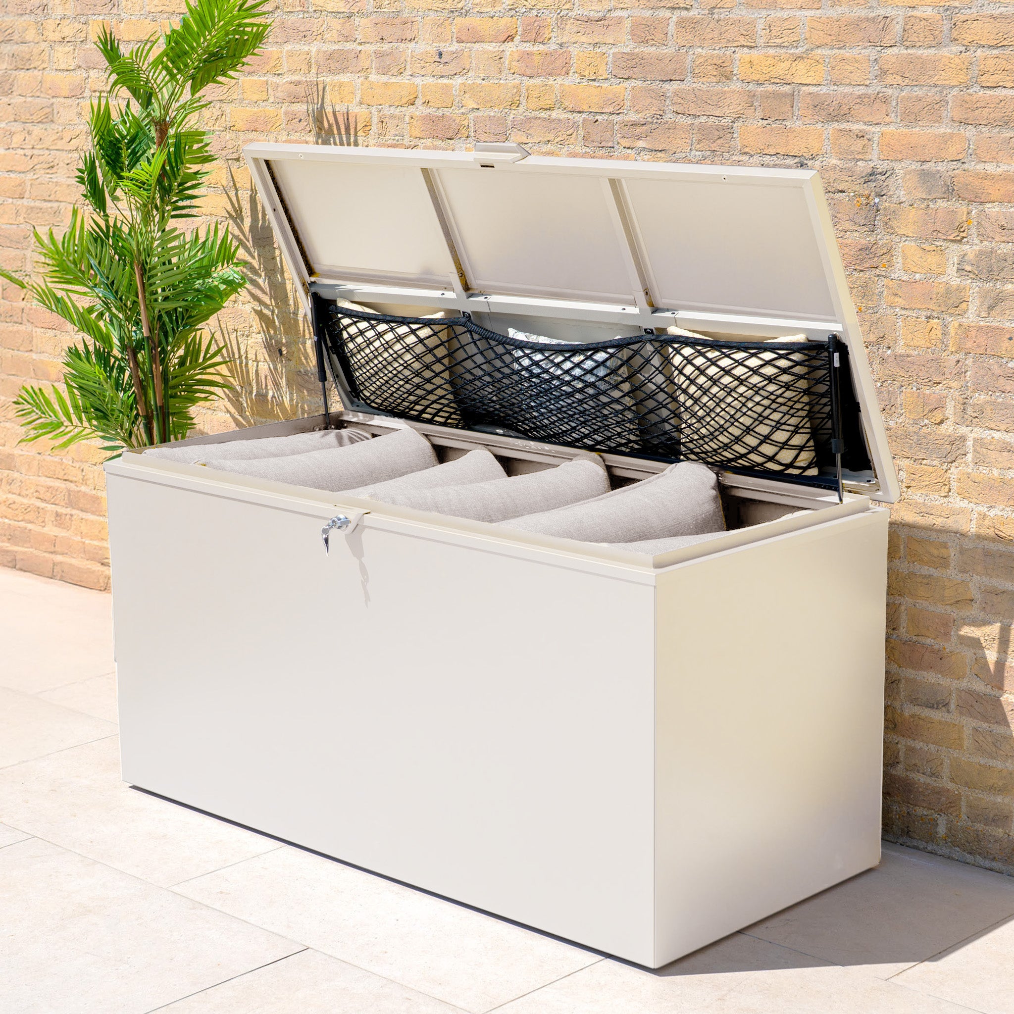 A Large Steel Storage Box in Latte with a watertight seal sits against a brick wall near a potted plant, its lid open to reveal outdoor storage for cushions.
