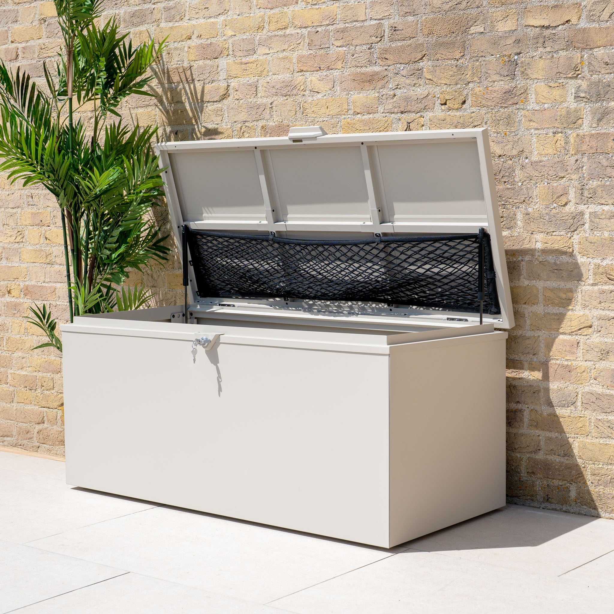 A Small Steel Storage Box in Latte with an open lid stands against a brick wall beside a potted green plant, perfect for outdoor storage.