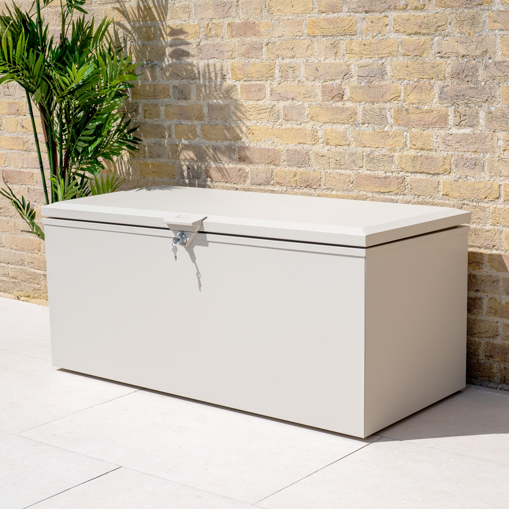The Small Steel Storage Box in Latte, shown by a brick wall and green potted plant, is perfect for keeping your outdoor items organized and secure.