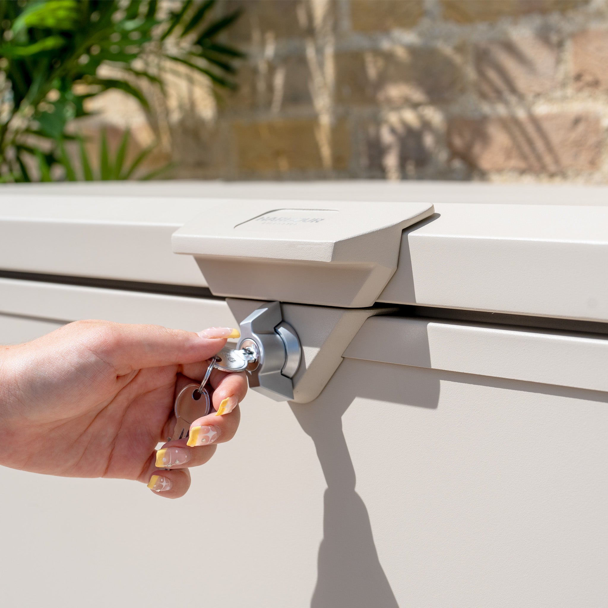 A hand uses a key to unlock the Small Steel Storage Box in Latte, shown outdoors with plants and a brick wall—ideal for keeping your items secure and dry.