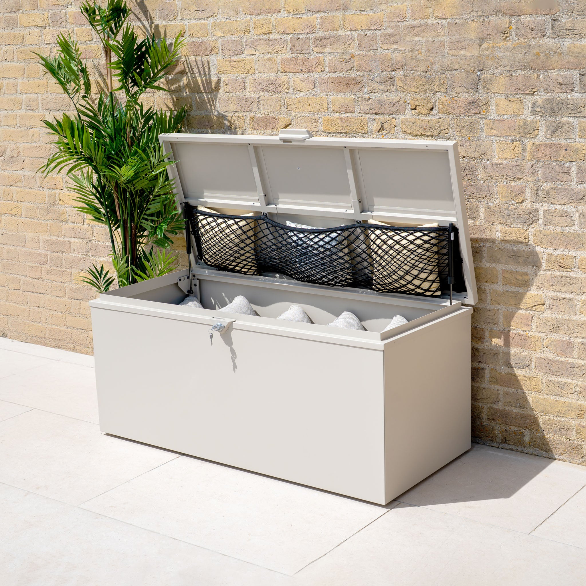 A Small Steel Storage Box in Latte with an open lid, offering watertight storage for pillows, sits beside a potted plant against a brick wall.
