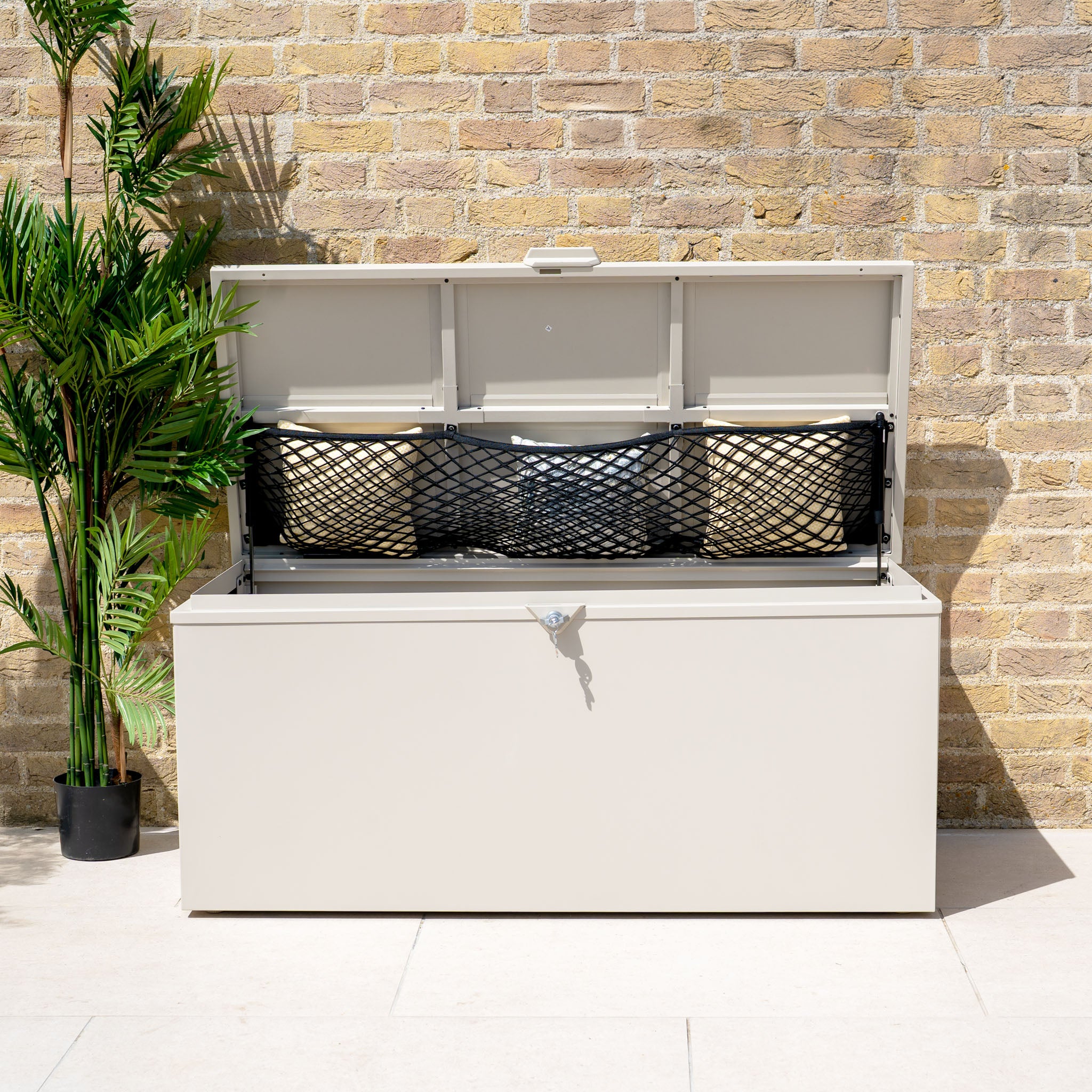 A Small Steel Storage Box in Latte with an open lid, black netting, and cushions inside sits against a brick wall—perfect for keeping patio essentials neat and dry.