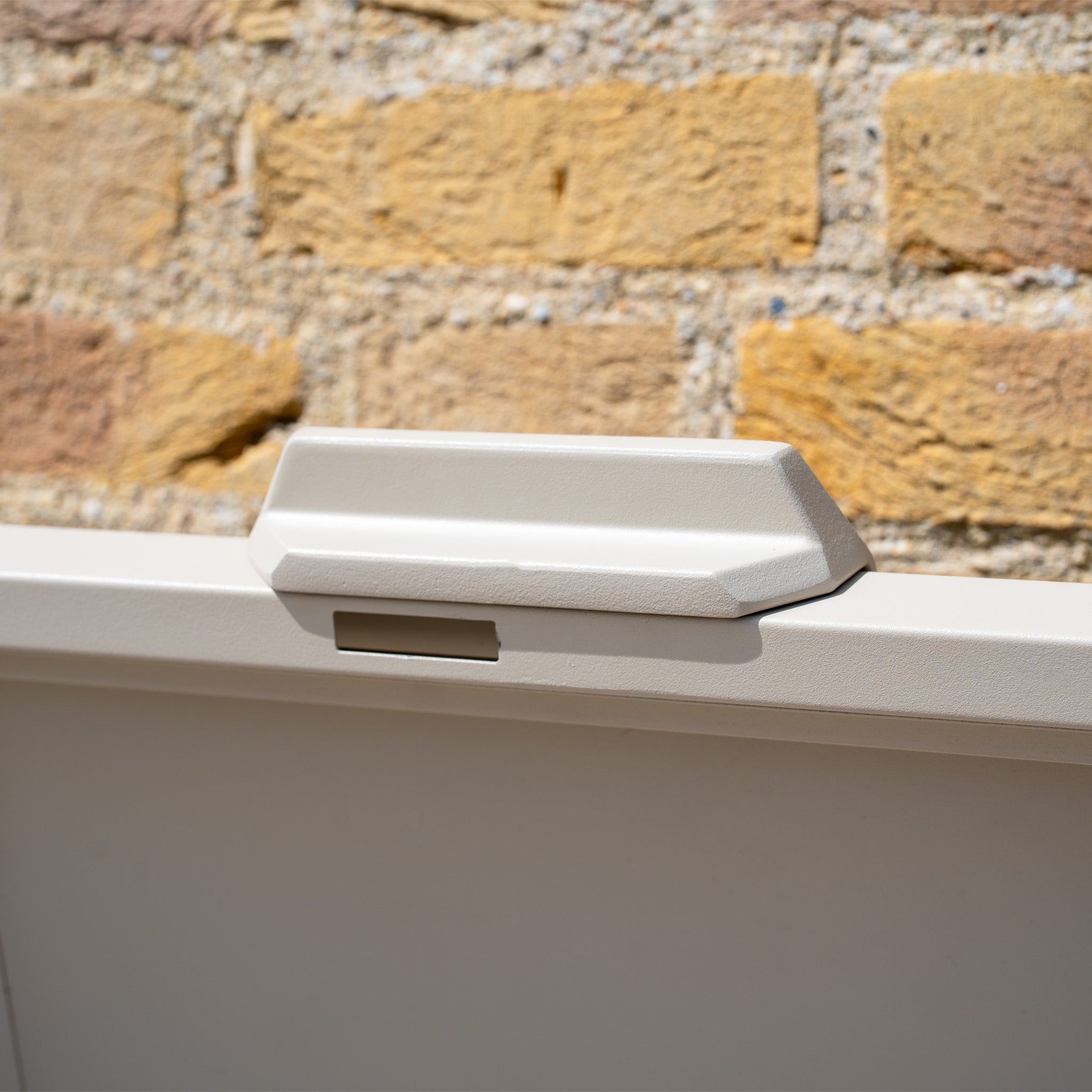 Close-up of the beige latch on the Large Steel Storage Box in Latte, with a brick wall visible behind the light-colored gate.