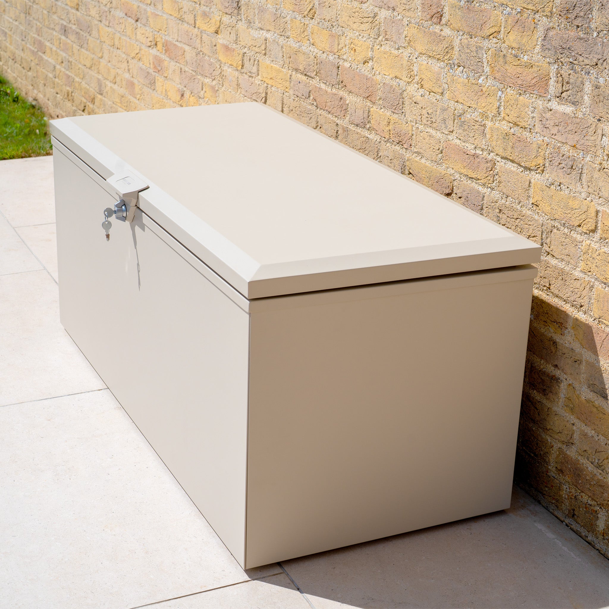 The Small Steel Storage Box in Latte, featuring a lock, sits on a patio by a brick wall—an ideal outdoor solution for keeping your items secure and organized.
