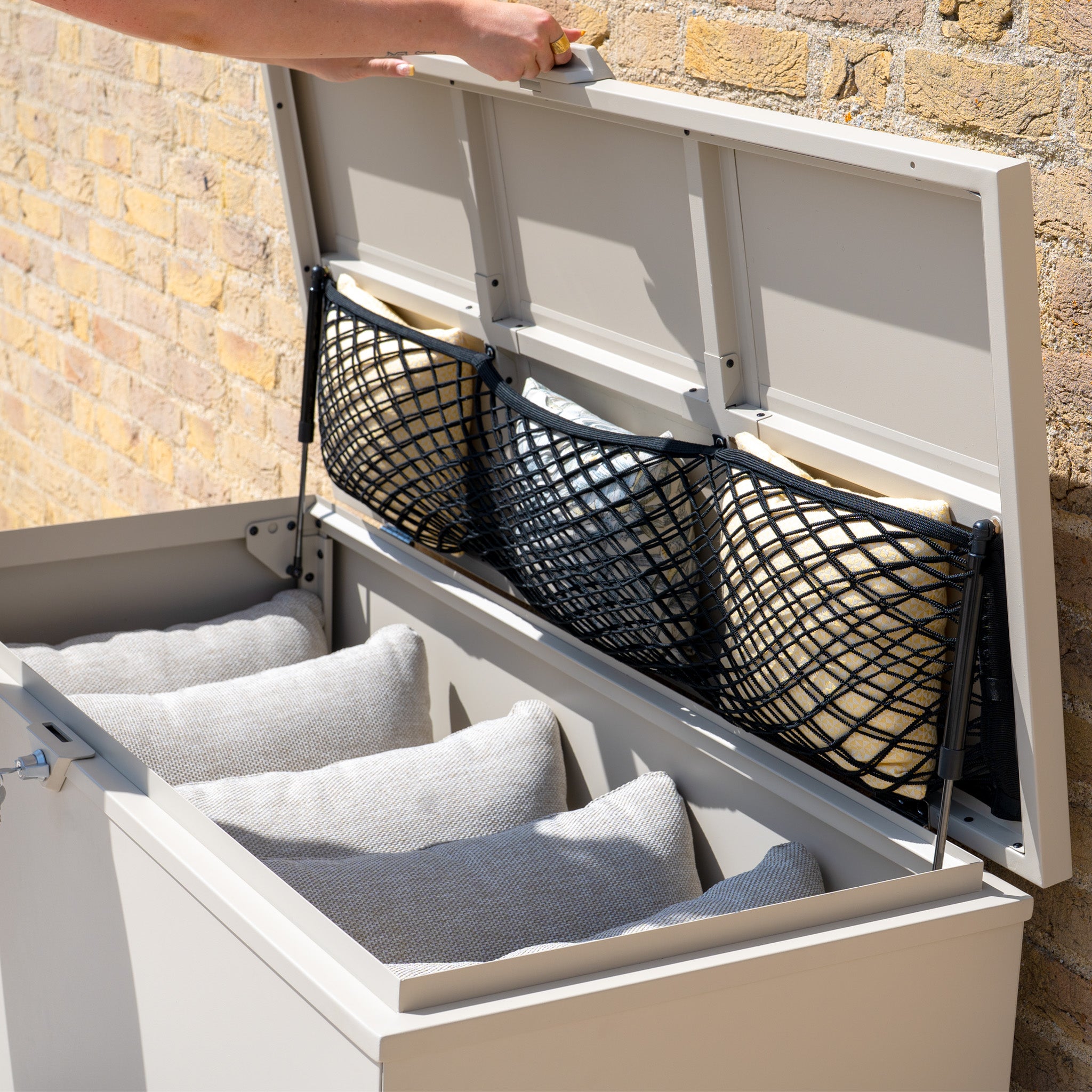 A person opens a Small Steel Storage Box in Latte, revealing cushions and a netted pocket inside the lid—perfect for organizing and protecting items in your outdoor space.