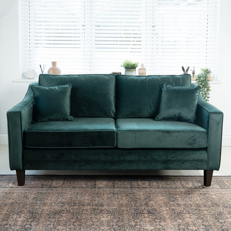 A Wiltshire 2 Seat Velvet Sofa in Bottle Green adds elegance to a bright room, complemented by two cushions, a window view, and lush plants.