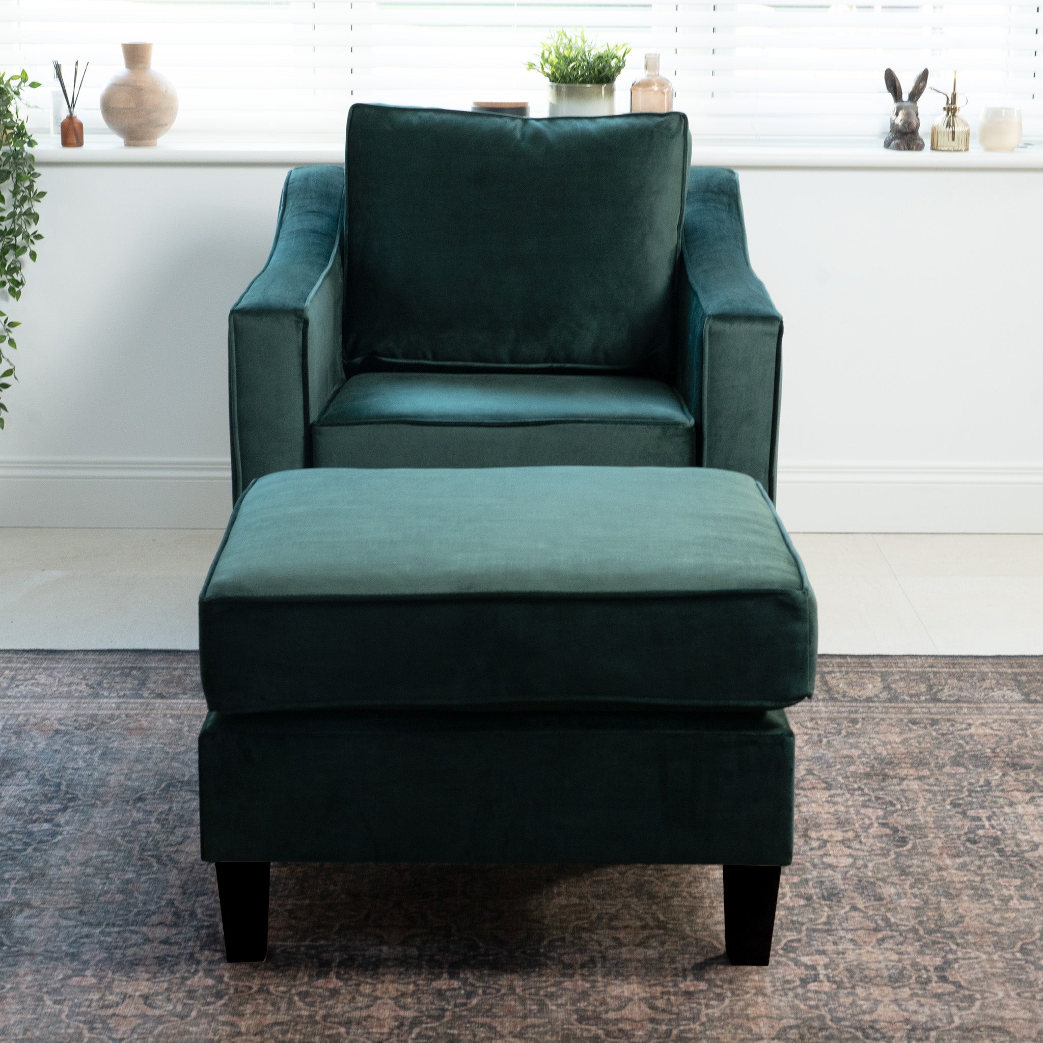 The Wiltshire Velvet Armchair in Bottle Green, paired with a matching ottoman, sits on a rug by a plant-adorned window. Handmade in England, it adds elegance and comfort to any room.