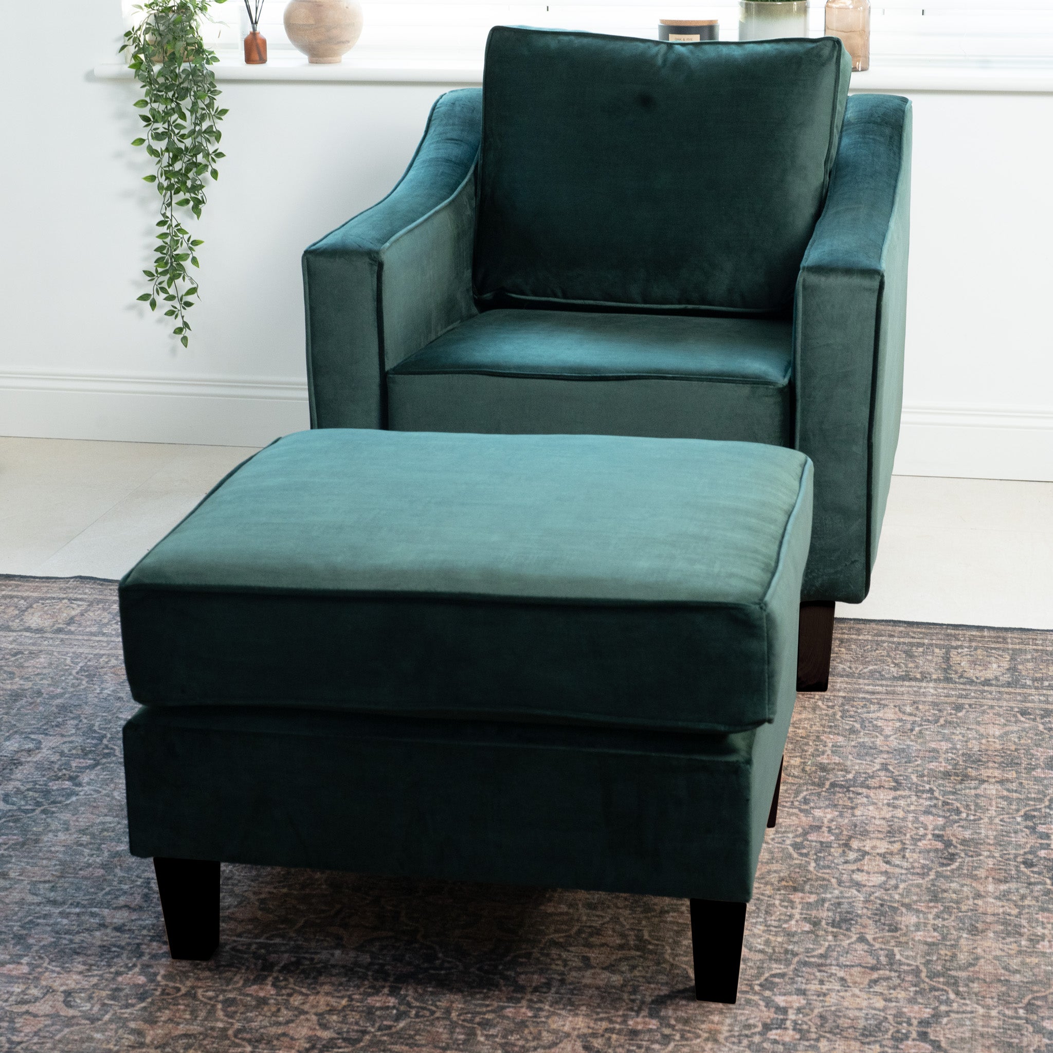 The bottle green Wiltshire Velvet Armchair is paired with a matching ottoman, sitting on a patterned rug in a well-lit room. Handmade in England, it exudes elegance and comfort.