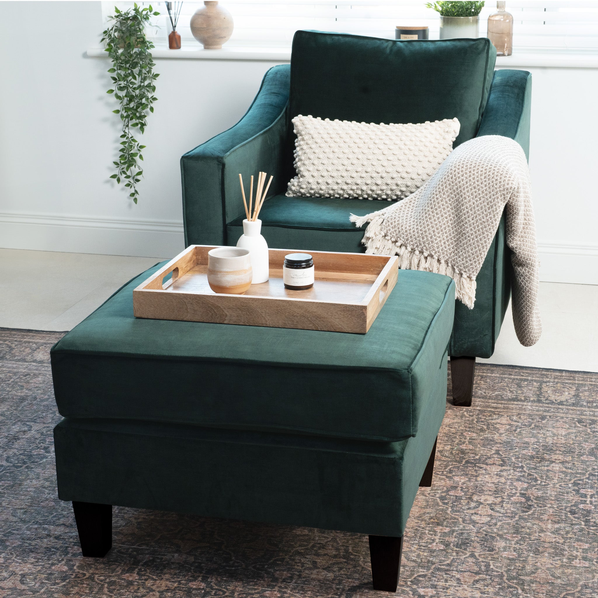 Handmade in England, the Wiltshire Velvet Armchair in rich bottle green offers warmth and style, complete with a cozy cushion and blanket. It elegantly pairs with an ottoman topped with a wooden tray and decor items.