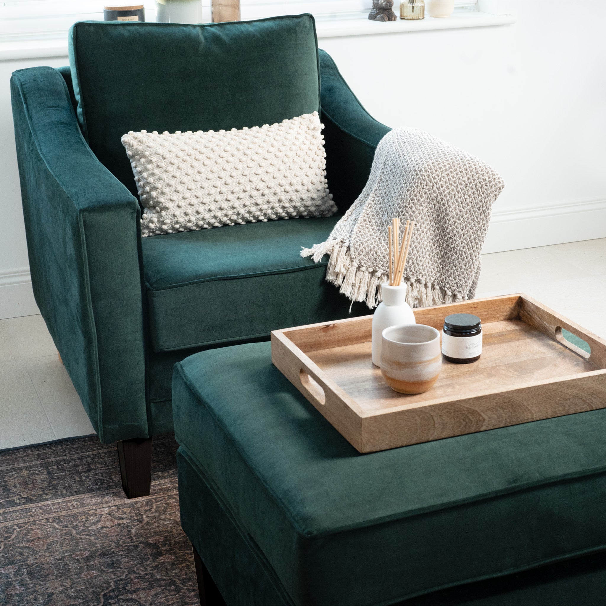 The handmade Wiltshire Velvet Armchair in Bottle Green, featuring a patterned pillow and blanket, stands elegantly next to a footrest that holds a wooden tray with decor items.
