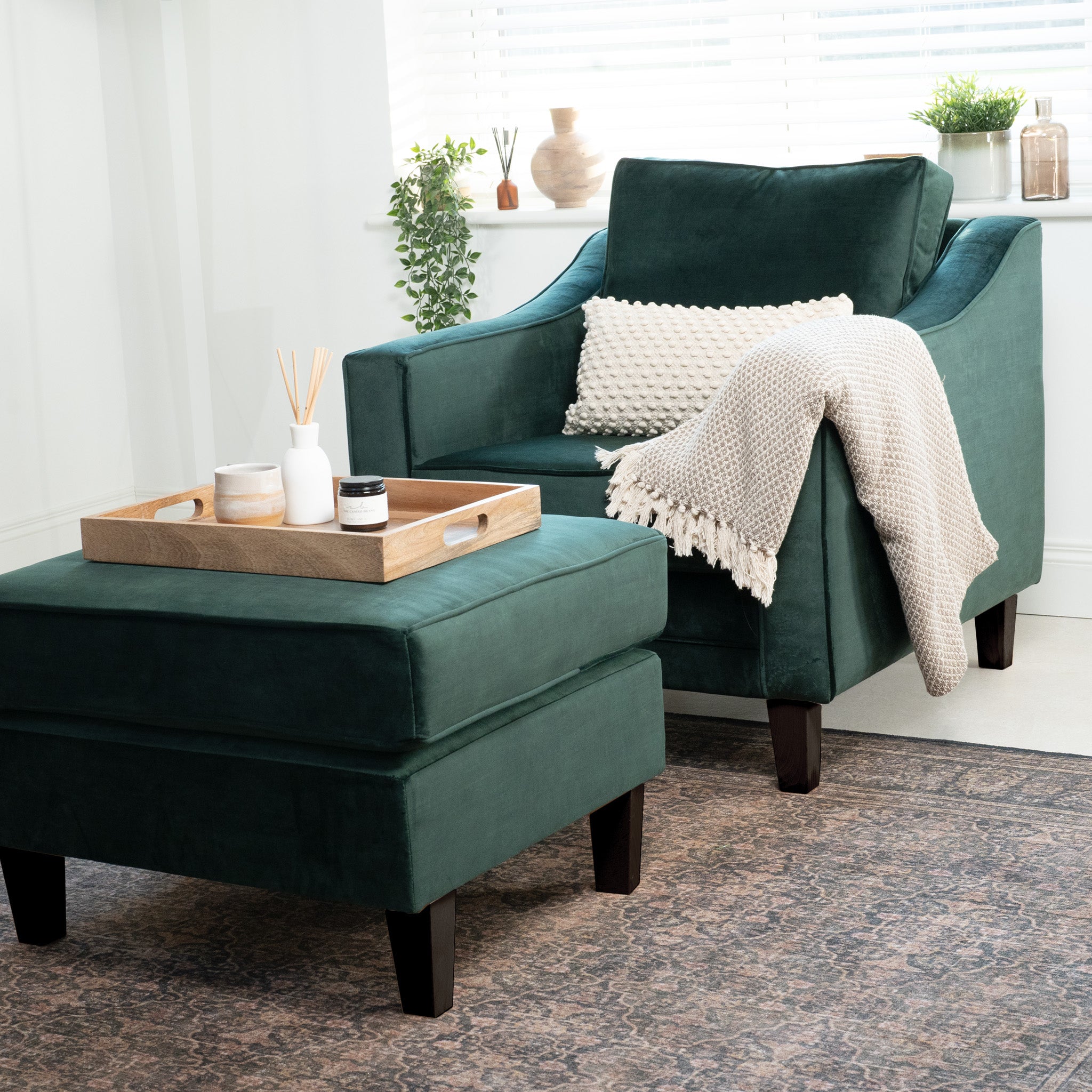 The Wiltshire Velvet Armchair in Bottle Green, featuring an ottoman, draped blanket, and a wooden tray on top, sits elegantly next to a window with blinds, adding warmth and style to any room.