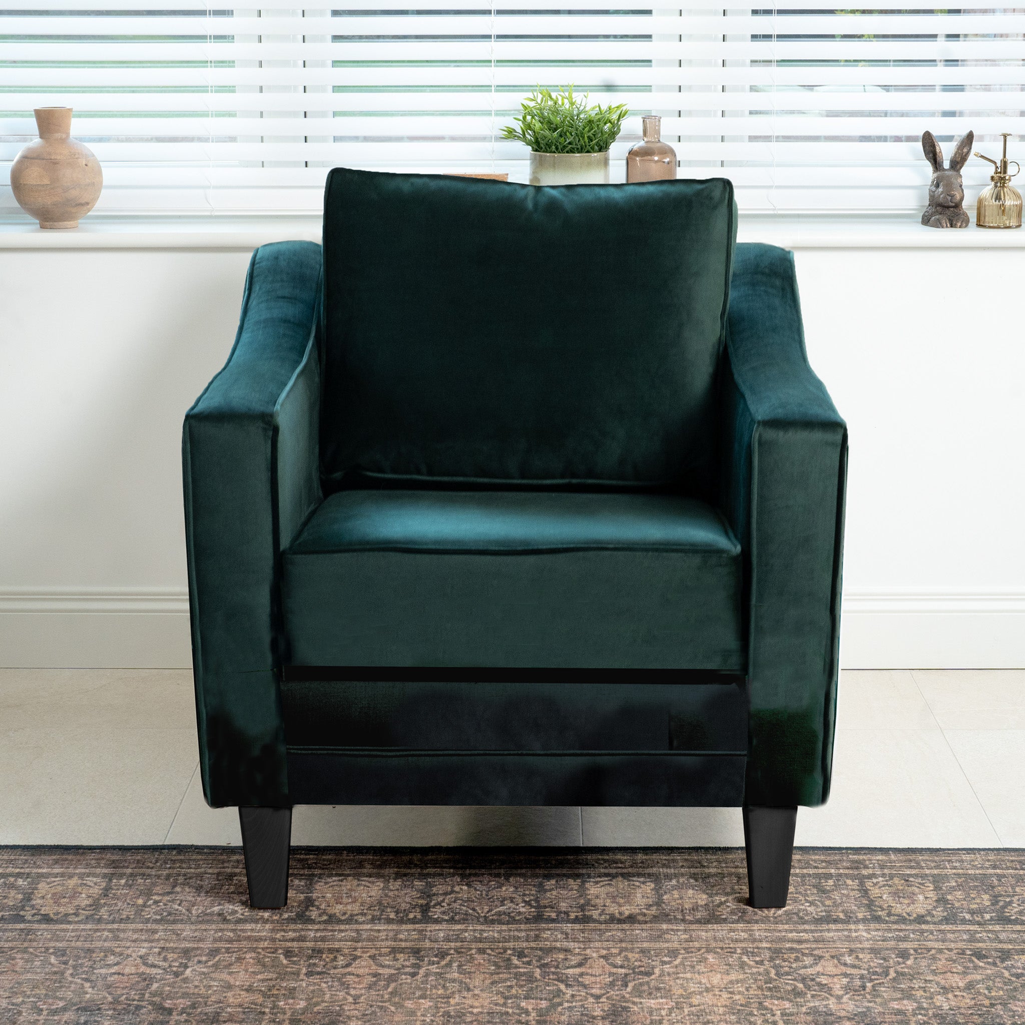 The Wiltshire Velvet Armchair in Bottle Green enhances the sunlit room with its handmade elegance, complemented by decorative windowsill items.
