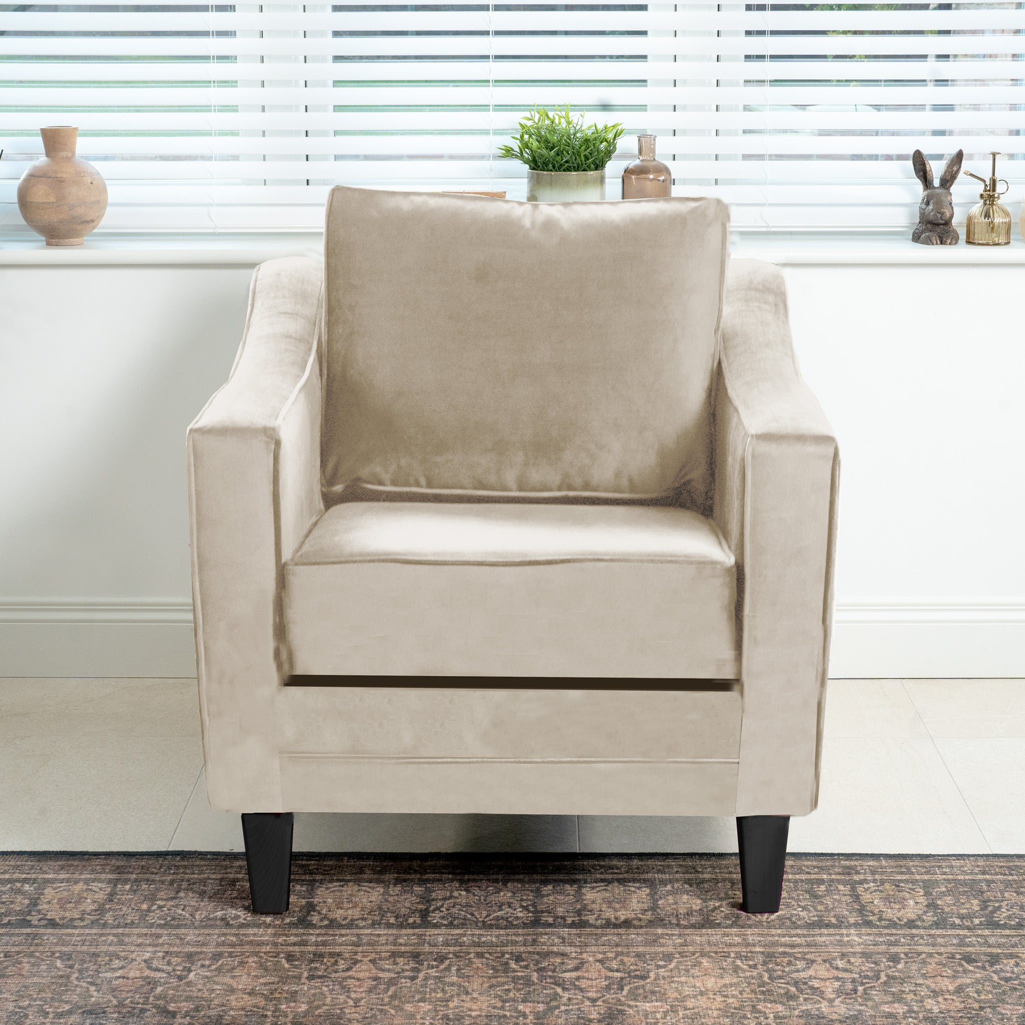 The Wiltshire Velvet Armchair in Cream takes center stage in the living room, with its soft texture enhanced by a rug and decorative window sill items. Handmade in England, this exquisite piece adds elegance to any space.