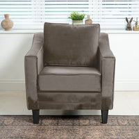 The Wiltshire Velvet Armchair in Mink, with elegant black legs, graces a bright room. Its luxurious velvet upholstery complements the nearby window adorned with blinds, plants, and decor. Handcrafted in England, this piece adds timeless charm to any setting.