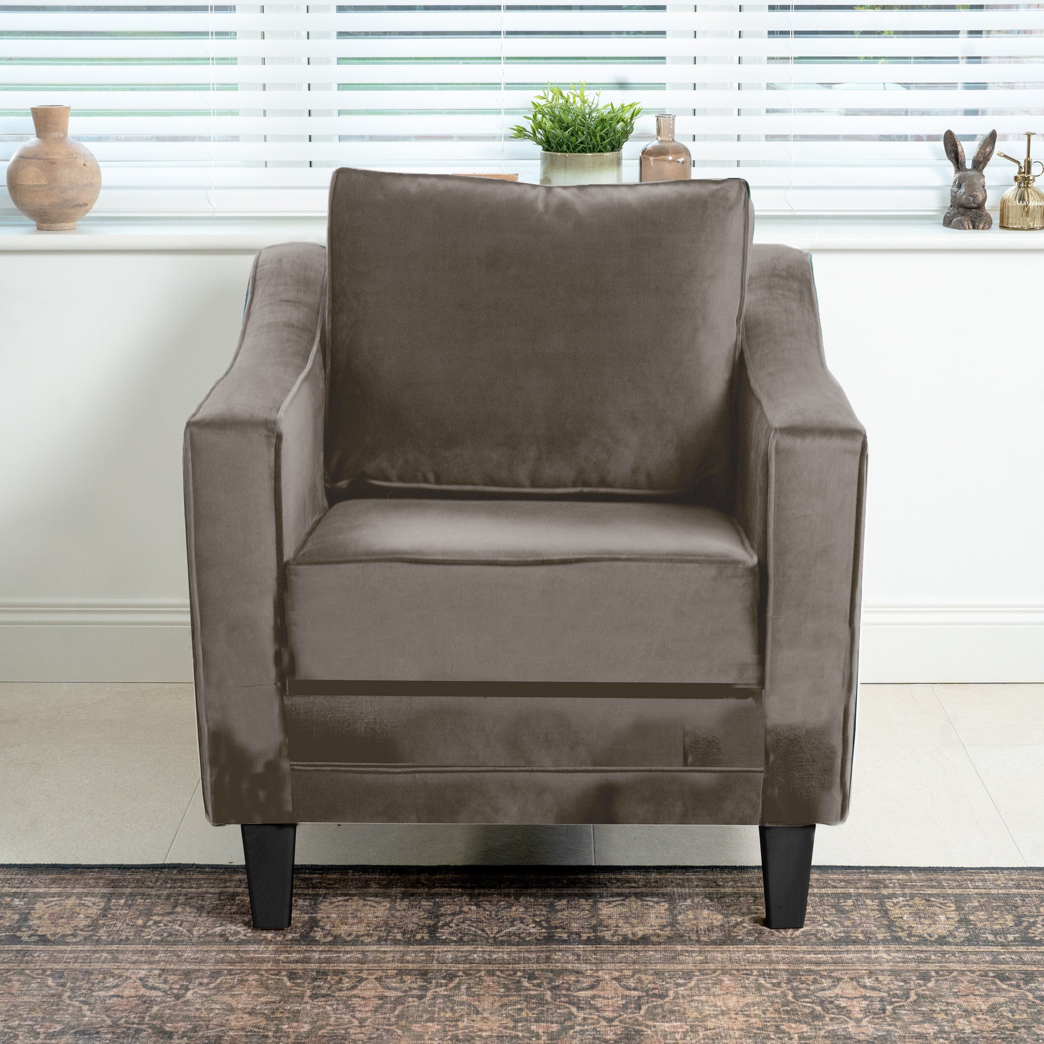 The Wiltshire Velvet Armchair in Mink, with elegant black legs, graces a bright room. Its luxurious velvet upholstery complements the nearby window adorned with blinds, plants, and decor. Handcrafted in England, this piece adds timeless charm to any setting.