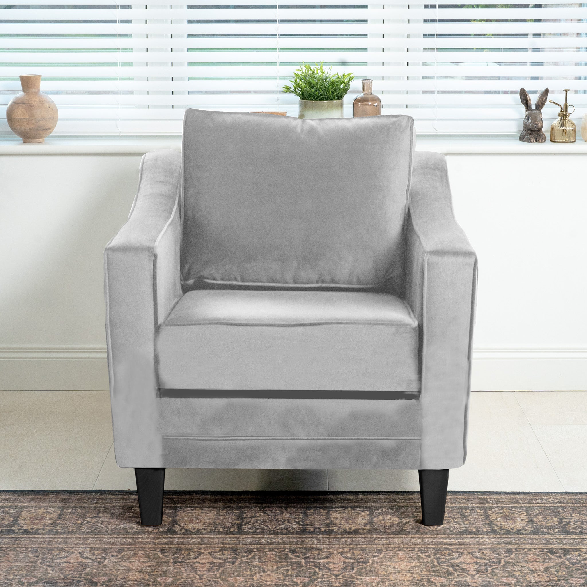 The Wiltshire Velvet Armchair in Feather Grey, with its square design, sits in front of the window beautifully adorned with plants and small sculptures on the sill.