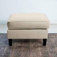 A Wiltshire Footstool in Beige with black legs elegantly rests on a patterned rug, adding modern flair to the space.