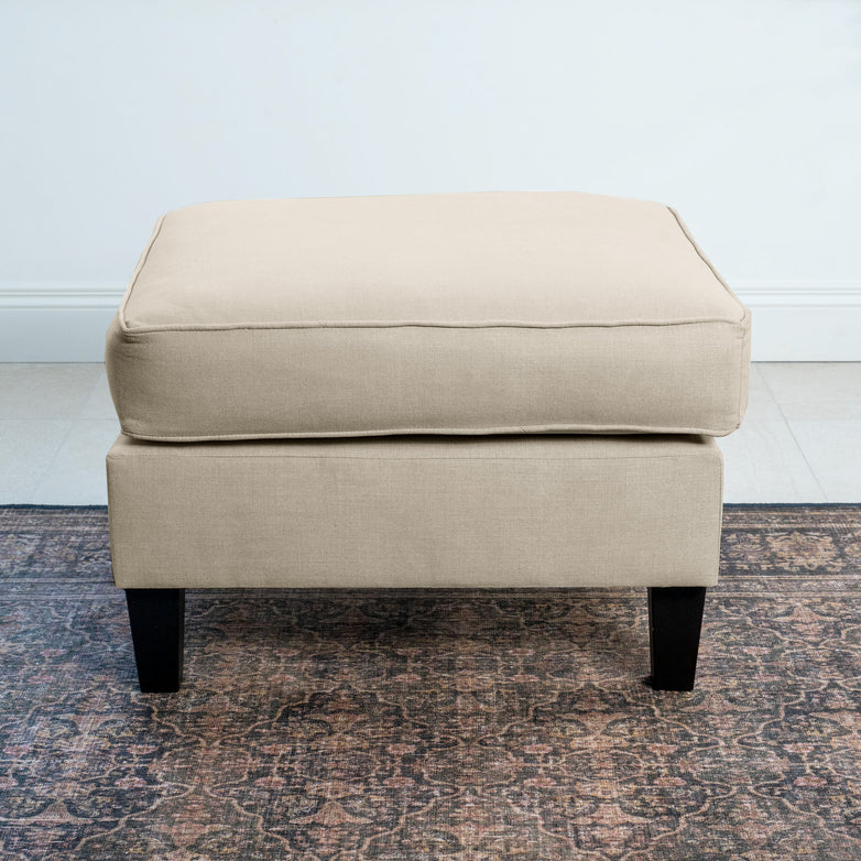 A Wiltshire Footstool in Beige with black legs elegantly rests on a patterned rug, adding modern flair to the space.