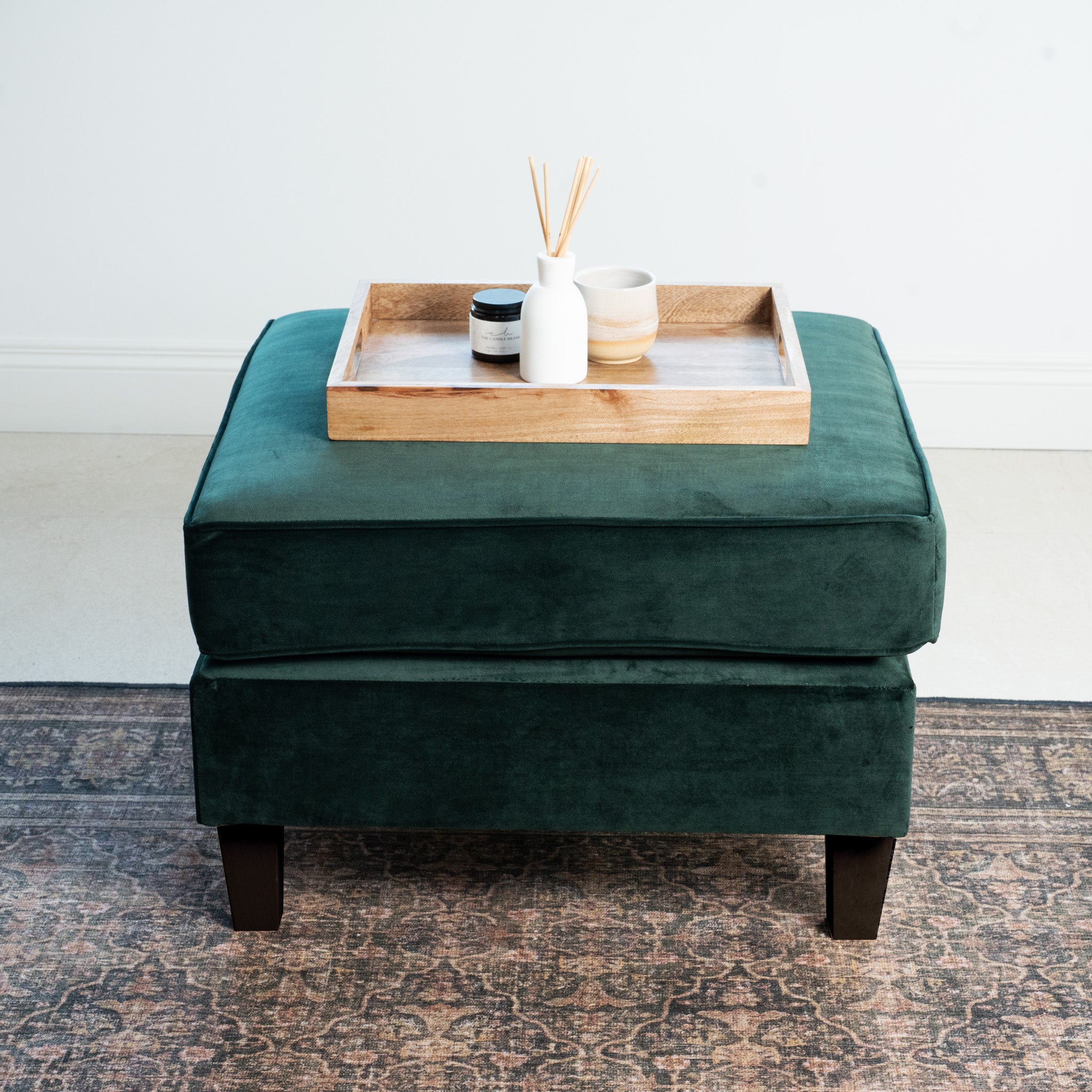 The Wiltshire Velvet Footstool in Bottle Green, a testament to handmade craftsmanship, elegantly holds a wooden tray with candles and a diffuser, all resting on a patterned rug.