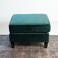 The Wiltshire Velvet Footstool in Bottle Green, featuring black legs, elegantly rests on a patterned carpet, enhancing the room's white walls and showcasing exquisite handmade craftsmanship.