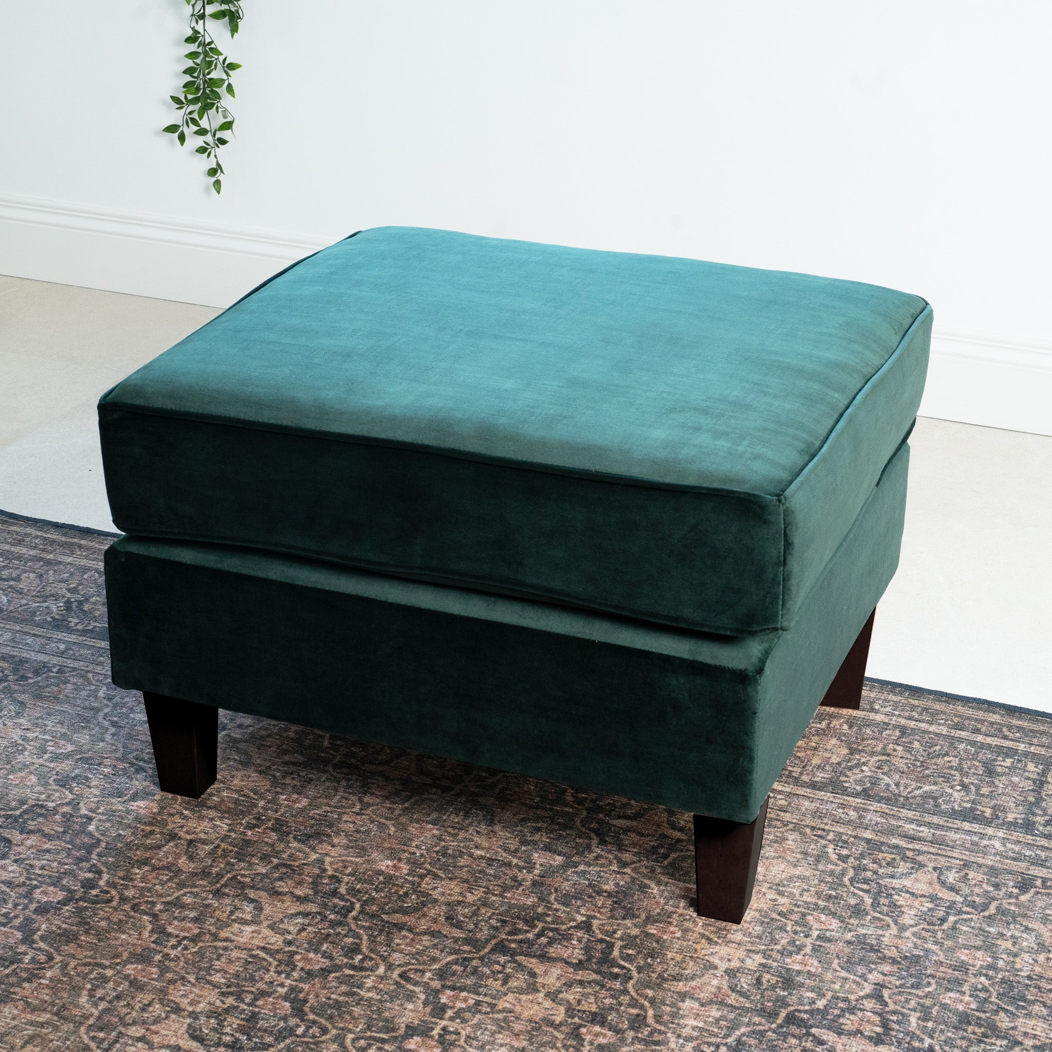 A Wiltshire Velvet Footstool in Bottle Green sits on a patterned rug in a bright room with light walls, complemented by the touch of handmade craftsmanship seen in the small plant decoration hanging nearby.