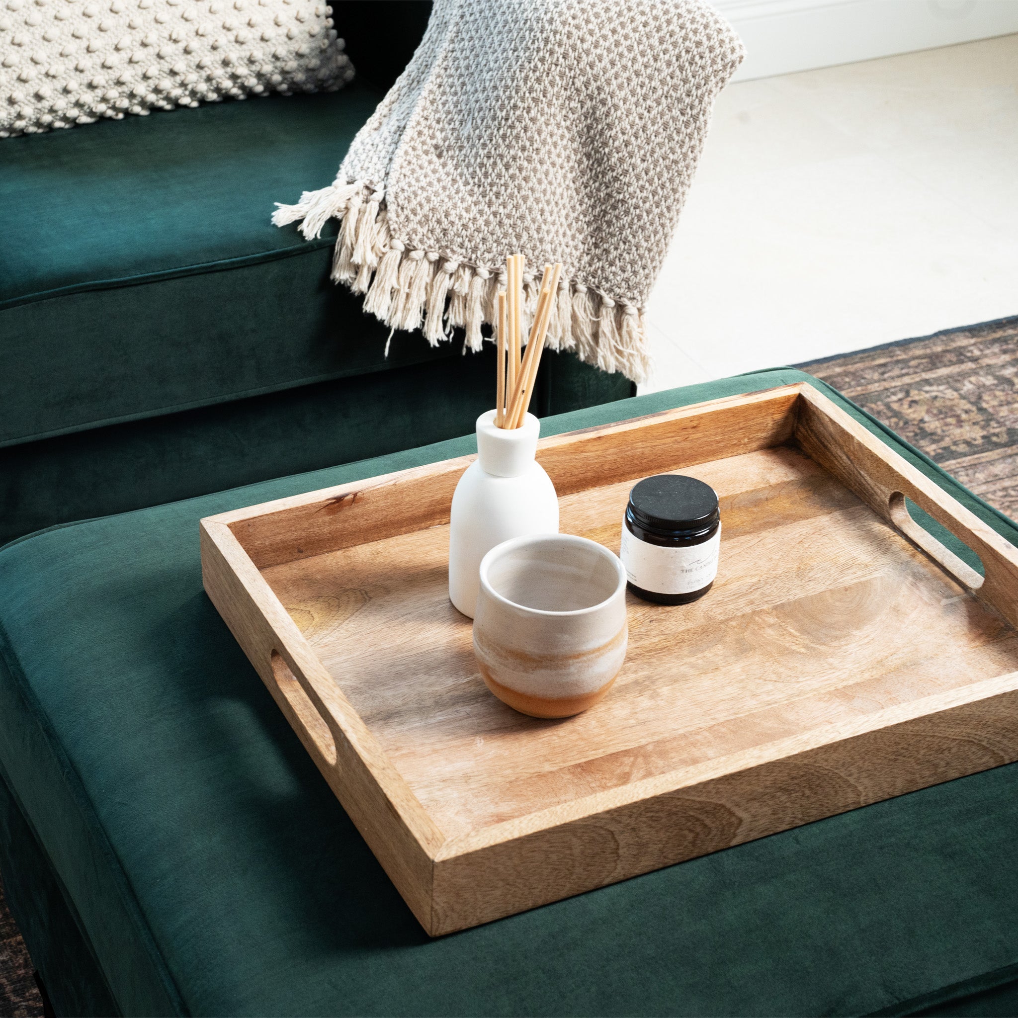 A Wiltshire Velvet Footstool in bottle green holds a wooden tray with a cup, diffuser, and jar, while a beige throw is draped over the ottoman.
