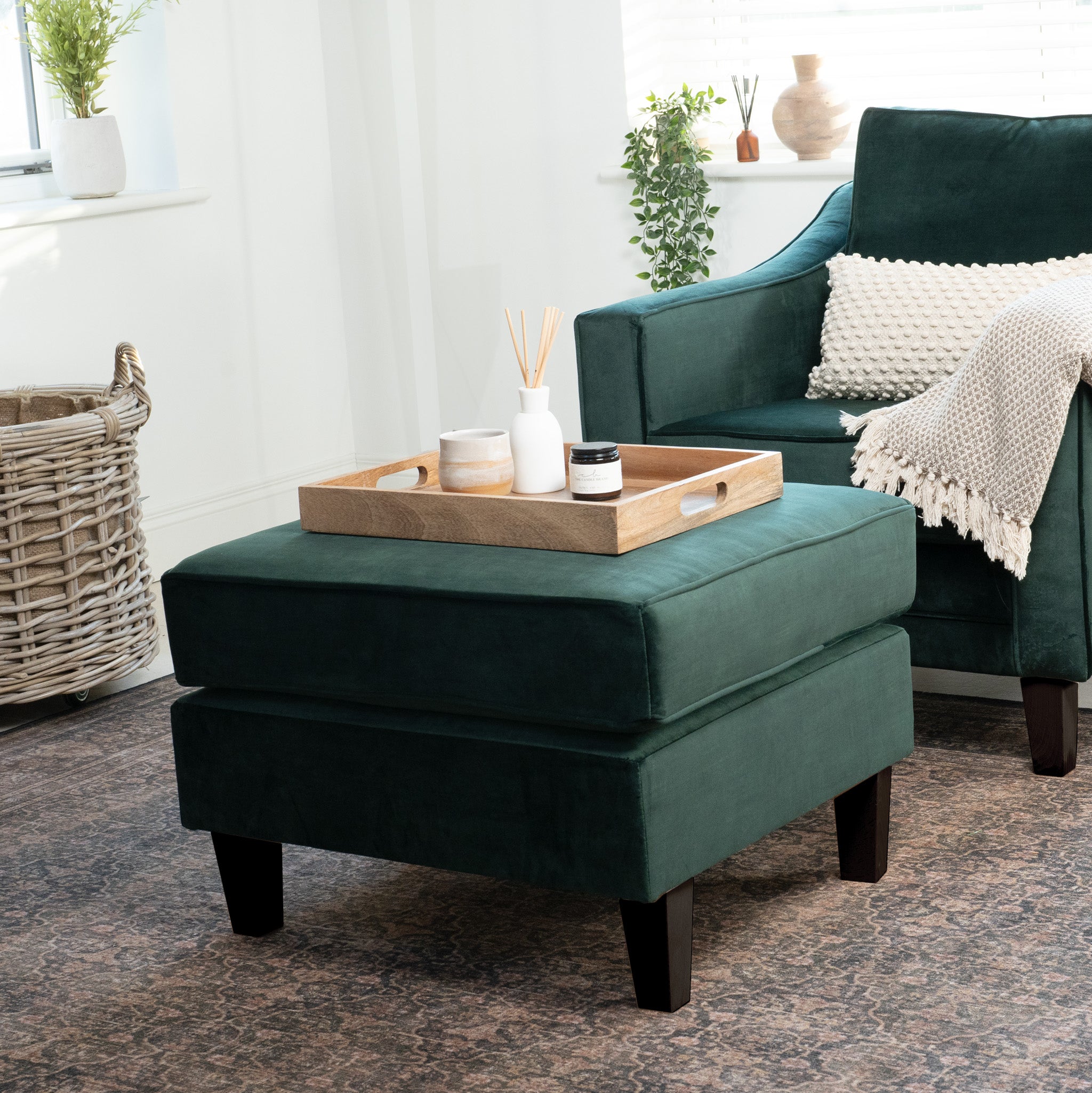 The Wiltshire Velvet Footstool in Bottle Green, featuring a wooden tray, sits next to a matching armchair in a cozy, sunlit living room.