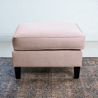 The Wiltshire Footstool in Dusky Pink, handcrafted, sits gracefully on a patterned rug in a minimalist room.