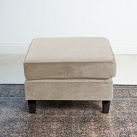 The Wiltshire Velvet Footstool in Cream, resting on a patterned rug with black legs, enhances the minimalist room, offering elegance and comfort.