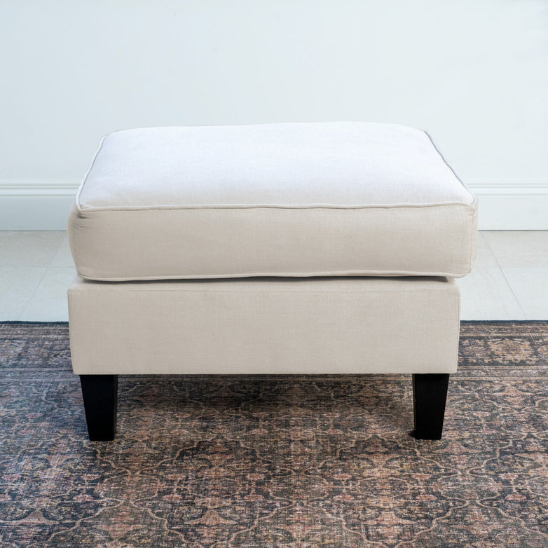 The Wiltshire Footstool in Ivory showcases ivory upholstery and a cushioned top, elegantly positioned on a patterned rug against a white wall.