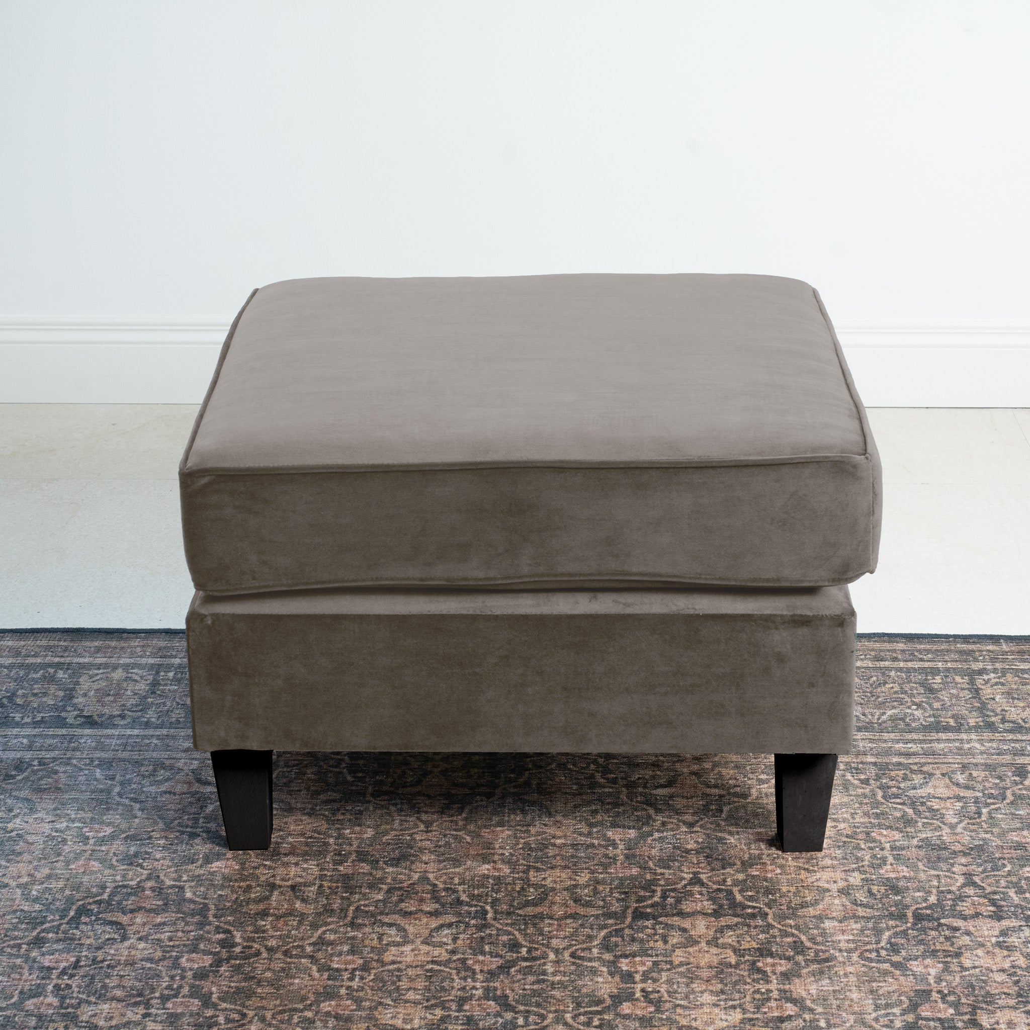 In a minimalist room, the Wiltshire Velvet Footstool in Mink with black legs sits elegantly on a patterned rug, showcasing superior craftsmanship.