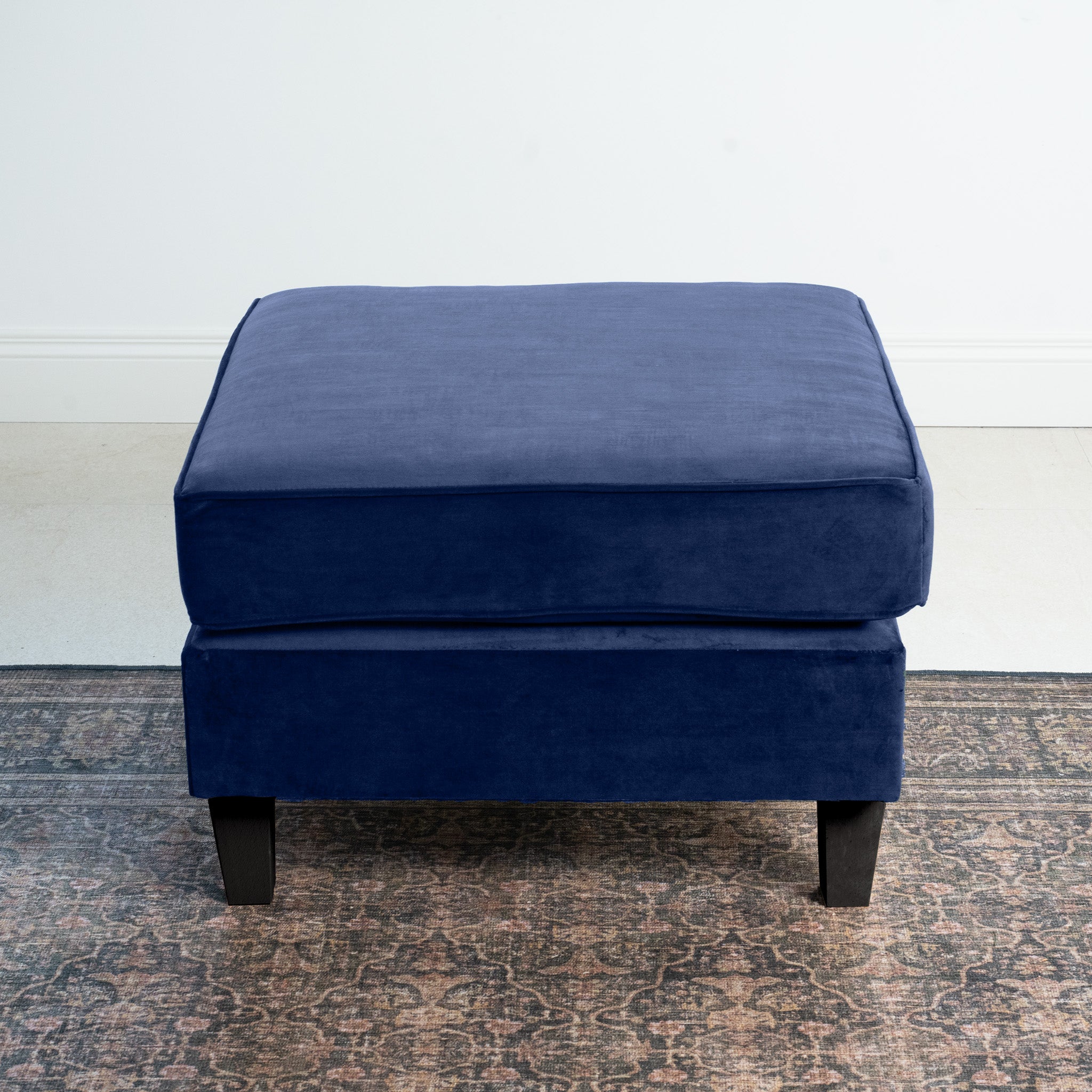 The Wiltshire Velvet Footstool in Royal Blue elegantly enhances the room on a patterned rug against a crisp white wall, adding contemporary flair.