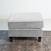 The Wiltshire Velvet Footstool in Feather Grey, crafted with timeless elegance, features sleek black legs and sits gracefully on a patterned rug in a minimalistic room.