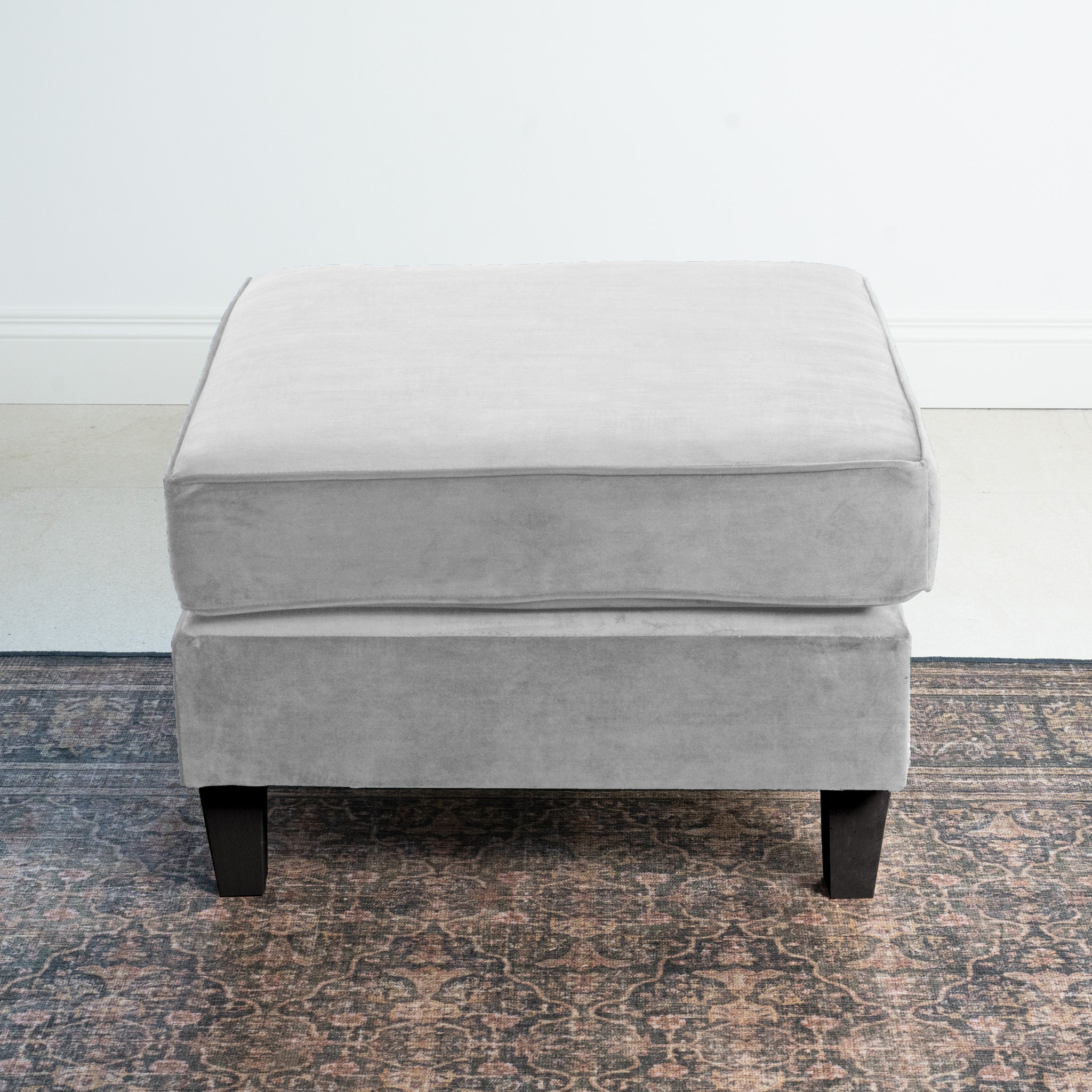 The Wiltshire Velvet Footstool in Feather Grey, crafted with timeless elegance, features sleek black legs and sits gracefully on a patterned rug in a minimalistic room.