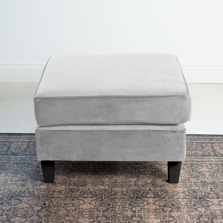 The Wiltshire Velvet Footstool in Feather Grey, crafted with timeless elegance, features sleek black legs and sits gracefully on a patterned rug in a minimalistic room.