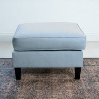 The Wiltshire Footstool in Sky Blue, featuring dark wooden legs, graces a patterned rug with elegance. Handcrafted with precision, this ottoman adds a touch of class to any space.