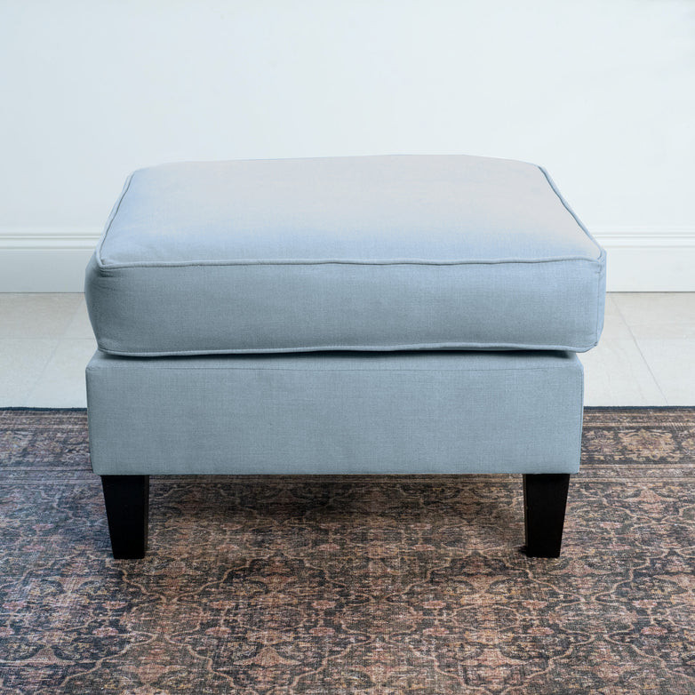 The Wiltshire Footstool in Sky Blue, featuring dark wooden legs, graces a patterned rug with elegance. Handcrafted with precision, this ottoman adds a touch of class to any space.